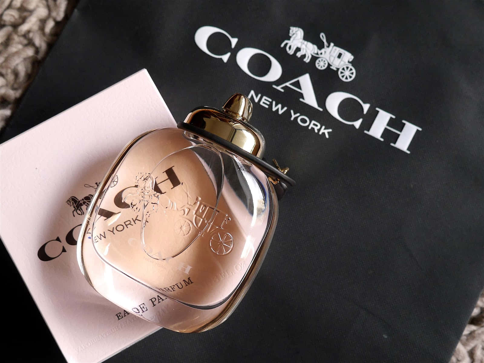 Coach 桌布 壁紙 — 一支玫瑰金色的Coach香水瓶優雅地放在其品牌包裝上，背景是帶有紋理的米色。