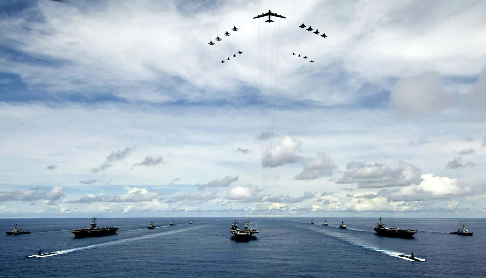Navy 桌布 壁紙 — 一支由航空母艦和軍艦組成的海軍艦隊，在陰天之下，戰鬥機在上空飛過，編隊航行在廣闊的藍色海洋上。