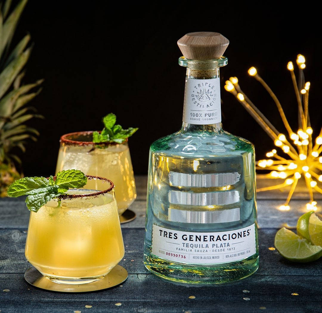 alcohol 桌布 壁紙 — 一支發光的 Tres Generaciones 龍舌蘭酒瓶放在一張深色木桌上，旁邊有兩杯以薄荷裝飾的雞尾酒，桌上還有溫暖的仙女燈和萊姆角。