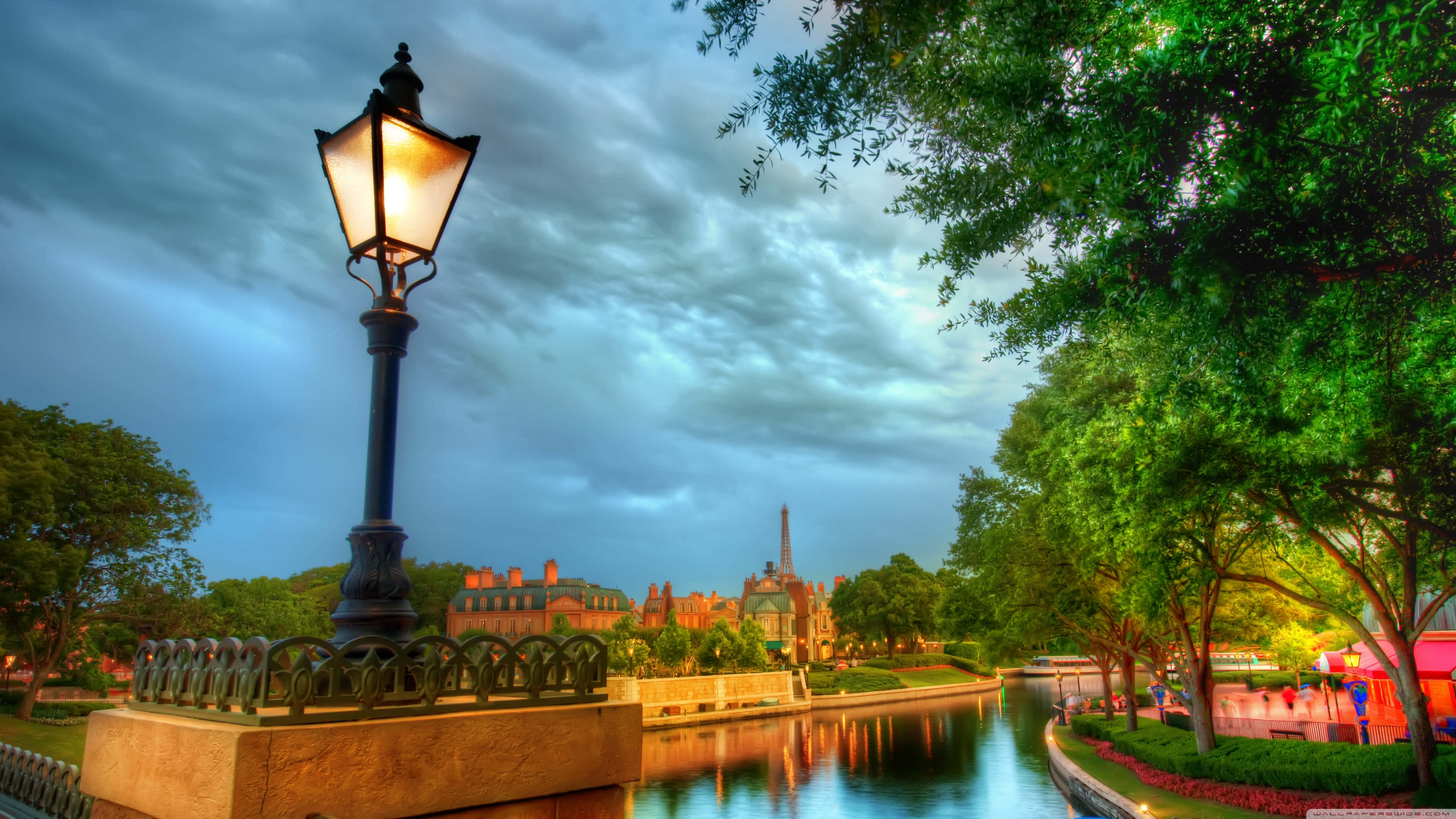 Disney World Desktop 桌布 壁紙 — 一支發光的復古街燈矗立在黃昏的運河旁，倒映在水中，樹木和遠處的建築物在陰沉天空下閃爍。