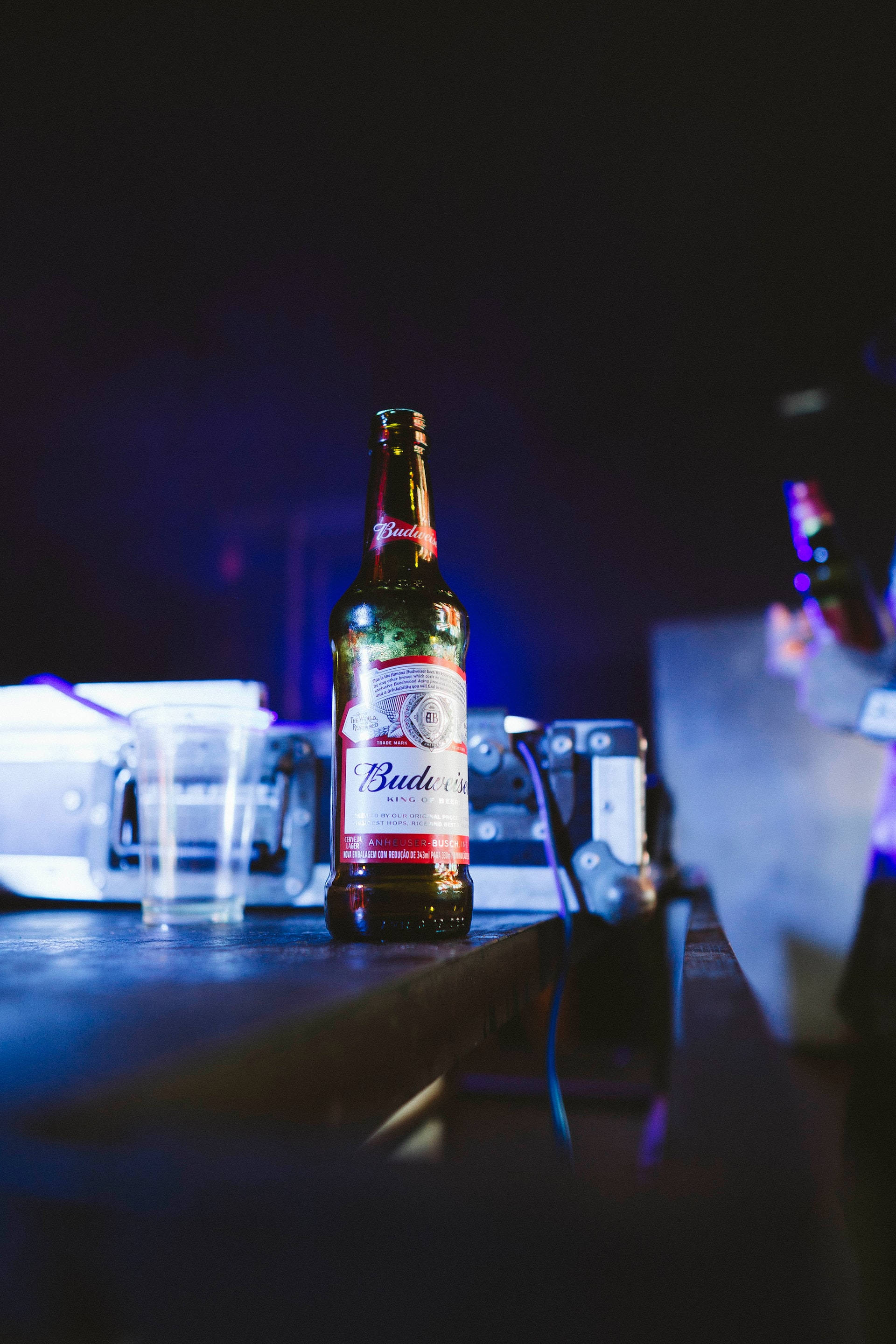 Drink 桌布 壁紙 — 一支百威啤酒瓶在霓虹燈下發光，背景是光線昏暗的酒吧檯面，上面有塑膠杯和設備。