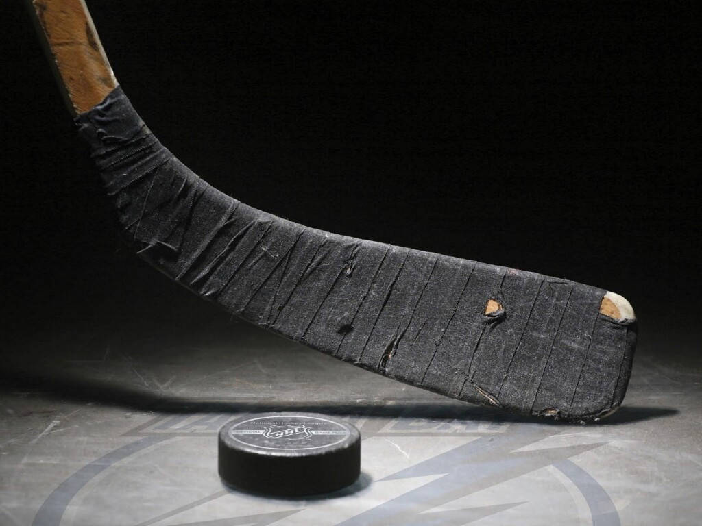 hockey 桌布 壁紙 — 一支磨損的曲棍球棍，帶有破損的膠帶，在戲劇性的聚光燈下傾斜在一個有刮痕的冰球場上的黑色冰球上方。