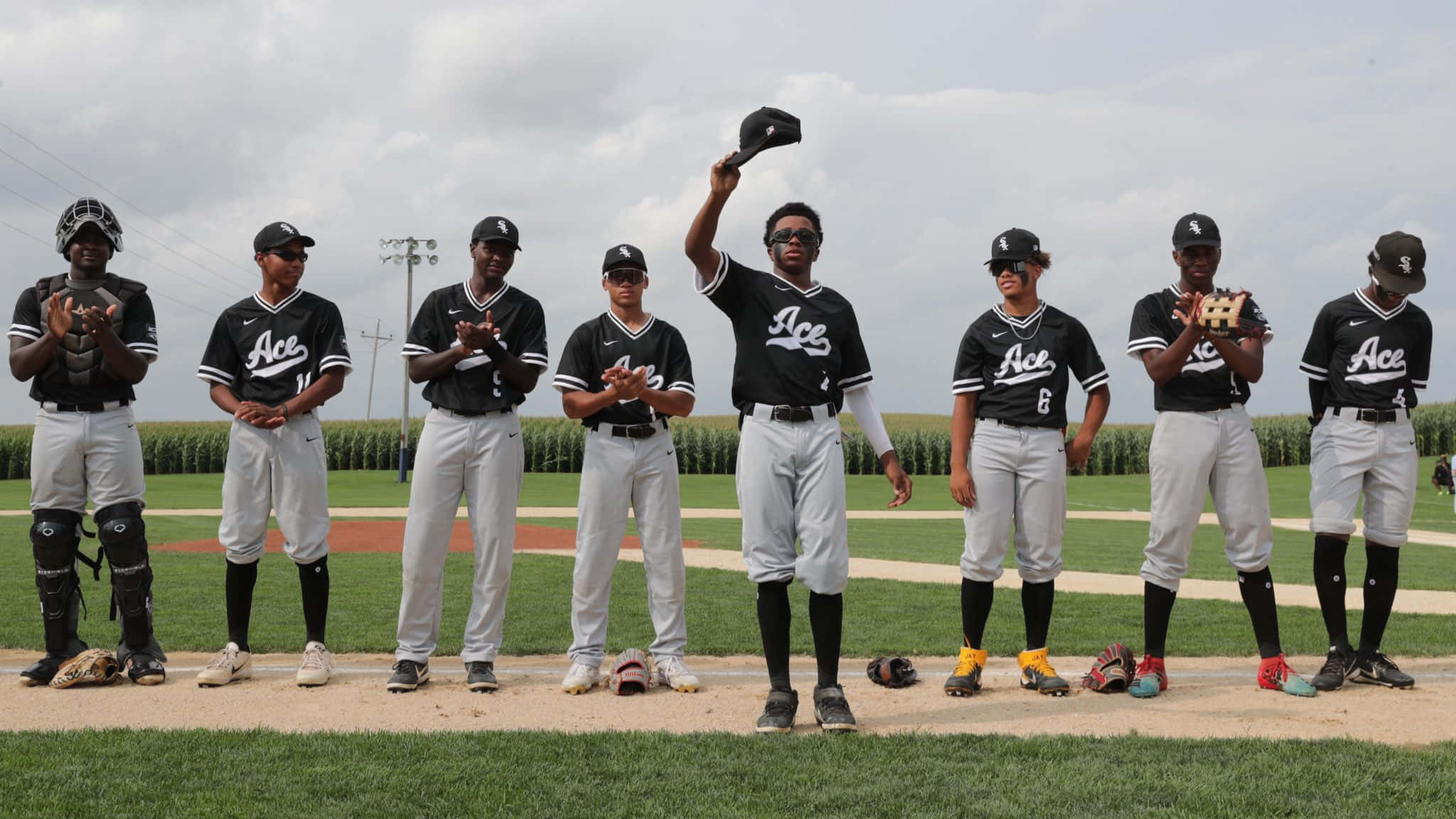 Little League Baseball 桌布 壁紙 — 一支穿著黑白球衣的棒球隊站在球場上，身後是玉米田，天空多雲。