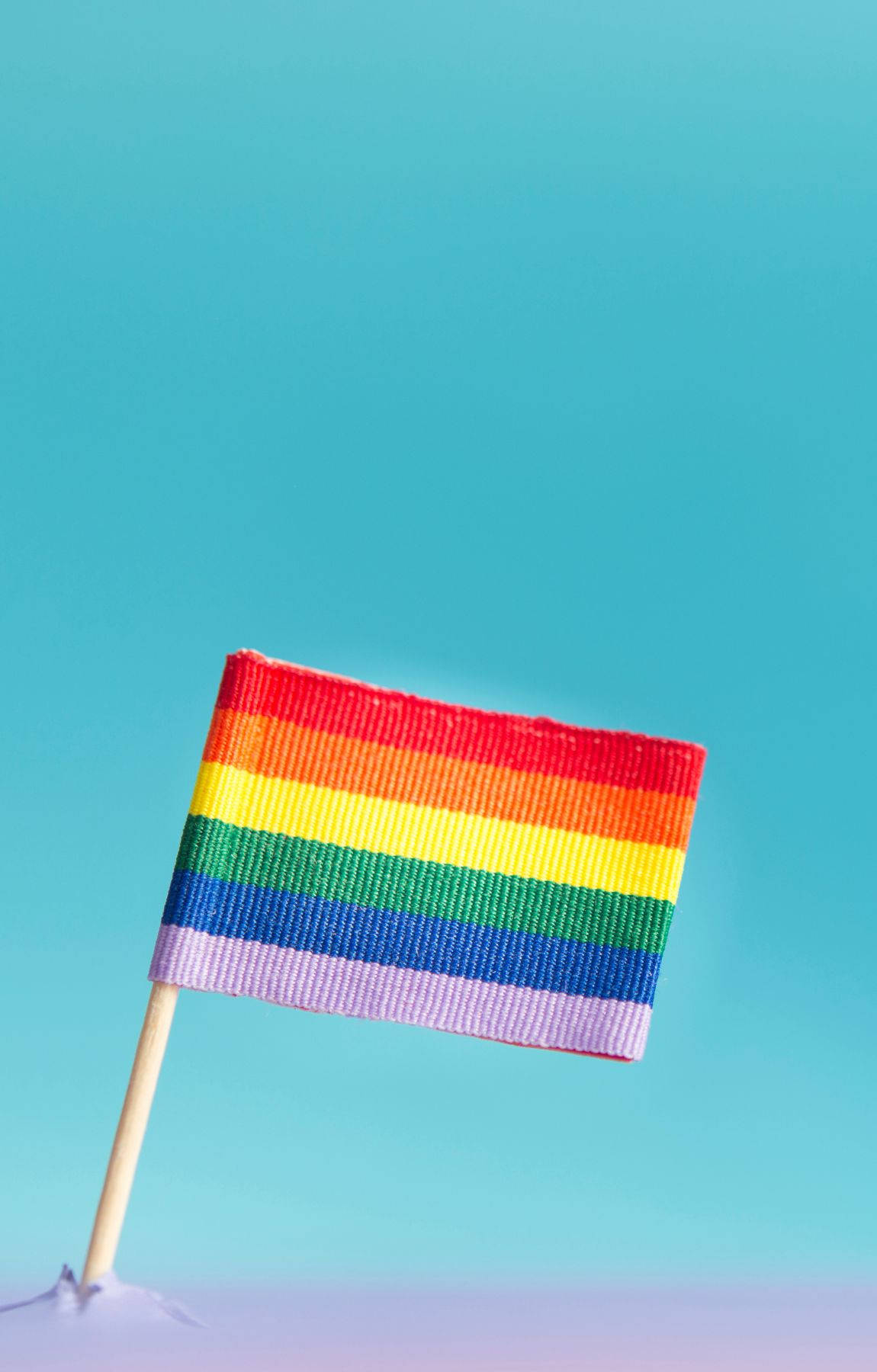Subtle Lgbt 桌布 壁紙 — 一支細小的彩虹旗插在棍子上，在柔和的藍色漸變背景前驕傲地豎立。