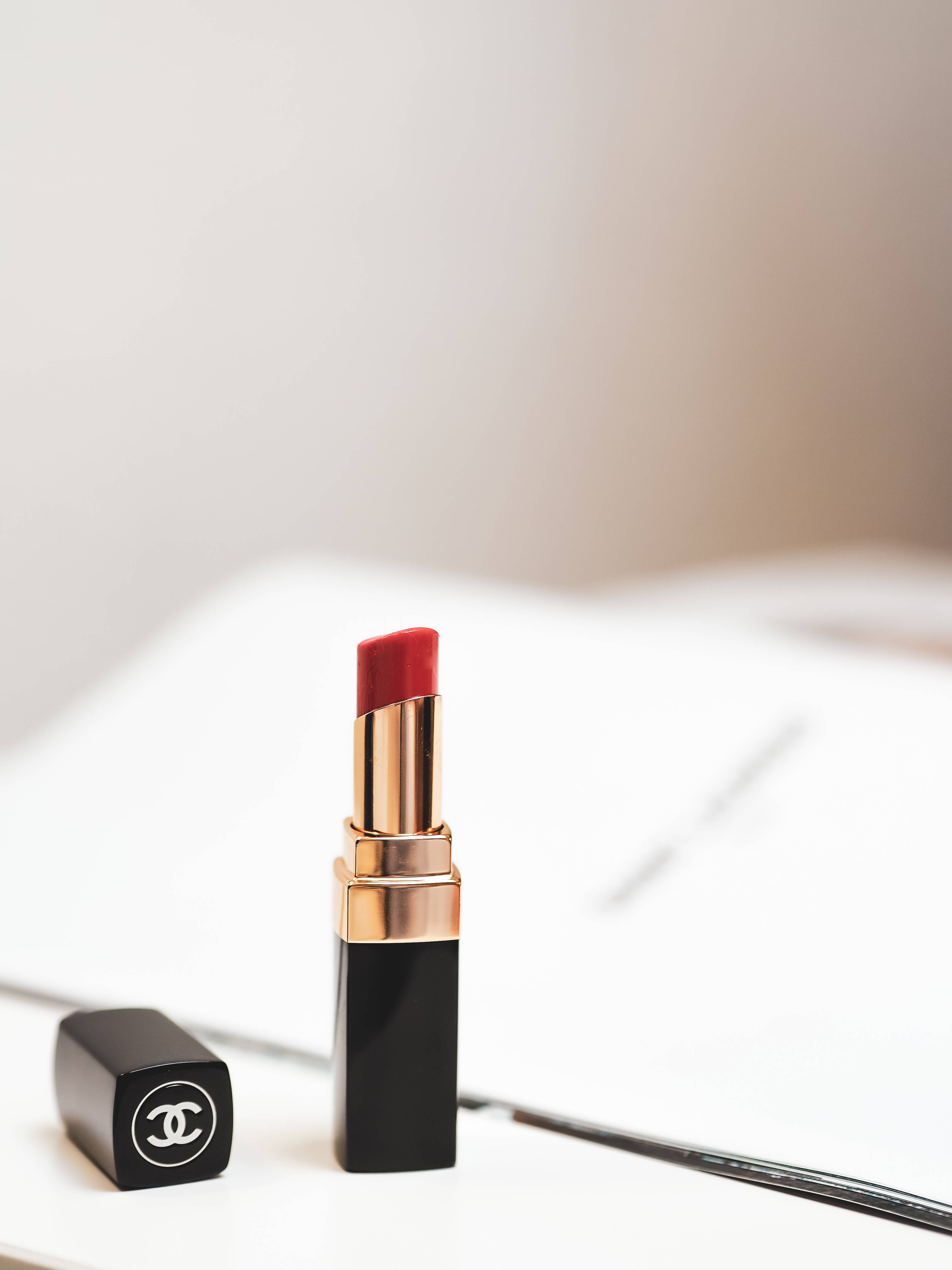 Chanel 桌布 壁紙 — 一支纖細的黑色Chanel唇膏，帶有金色點綴和紅色子彈，放在白色表面上，旁邊是其蓋子。
