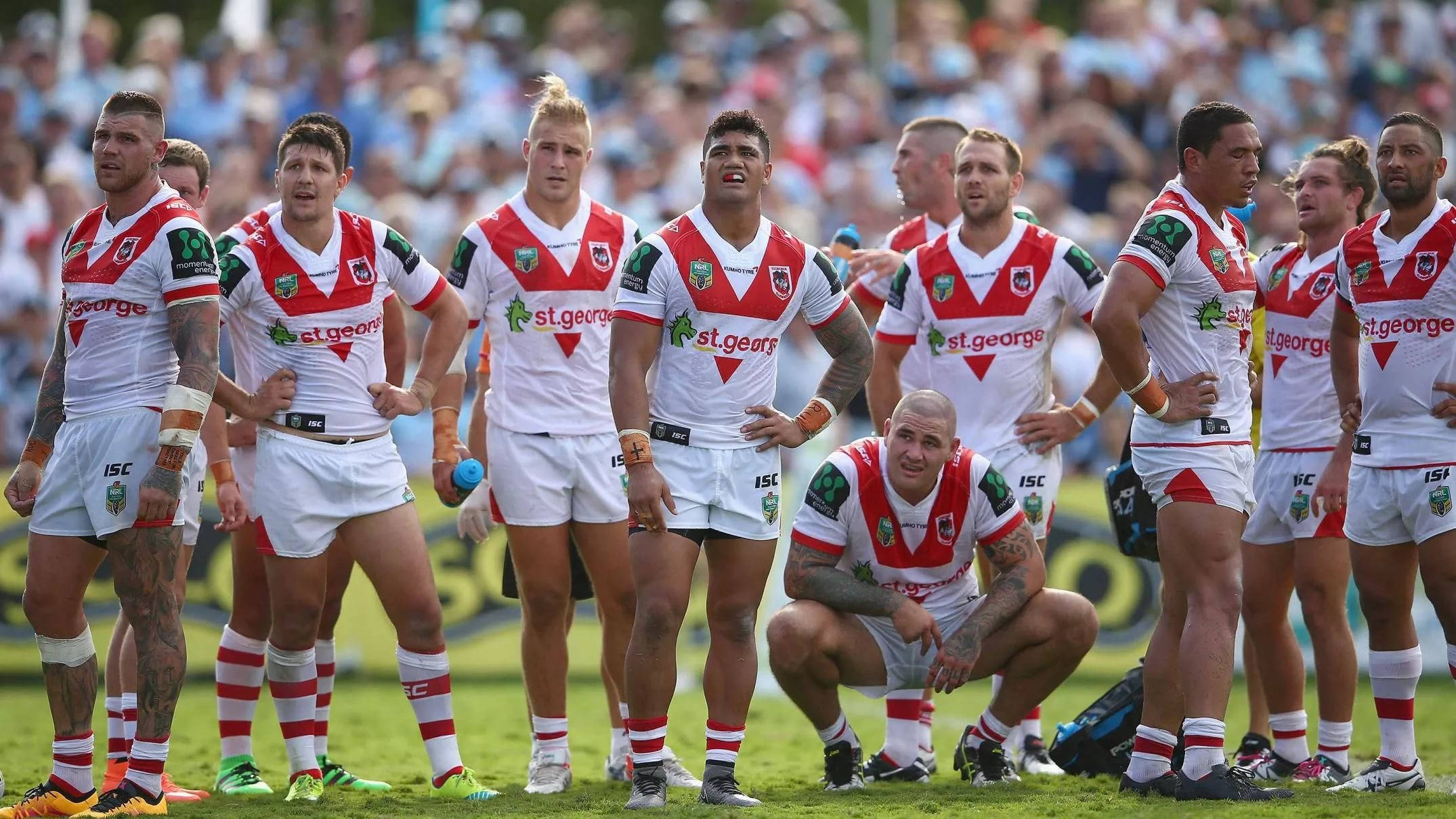 Nrl 桌布 壁紙 — 一支身穿紅白球衣的橄欖球聯賽隊伍在綠色球場上站立和跪著，背景有觀眾。