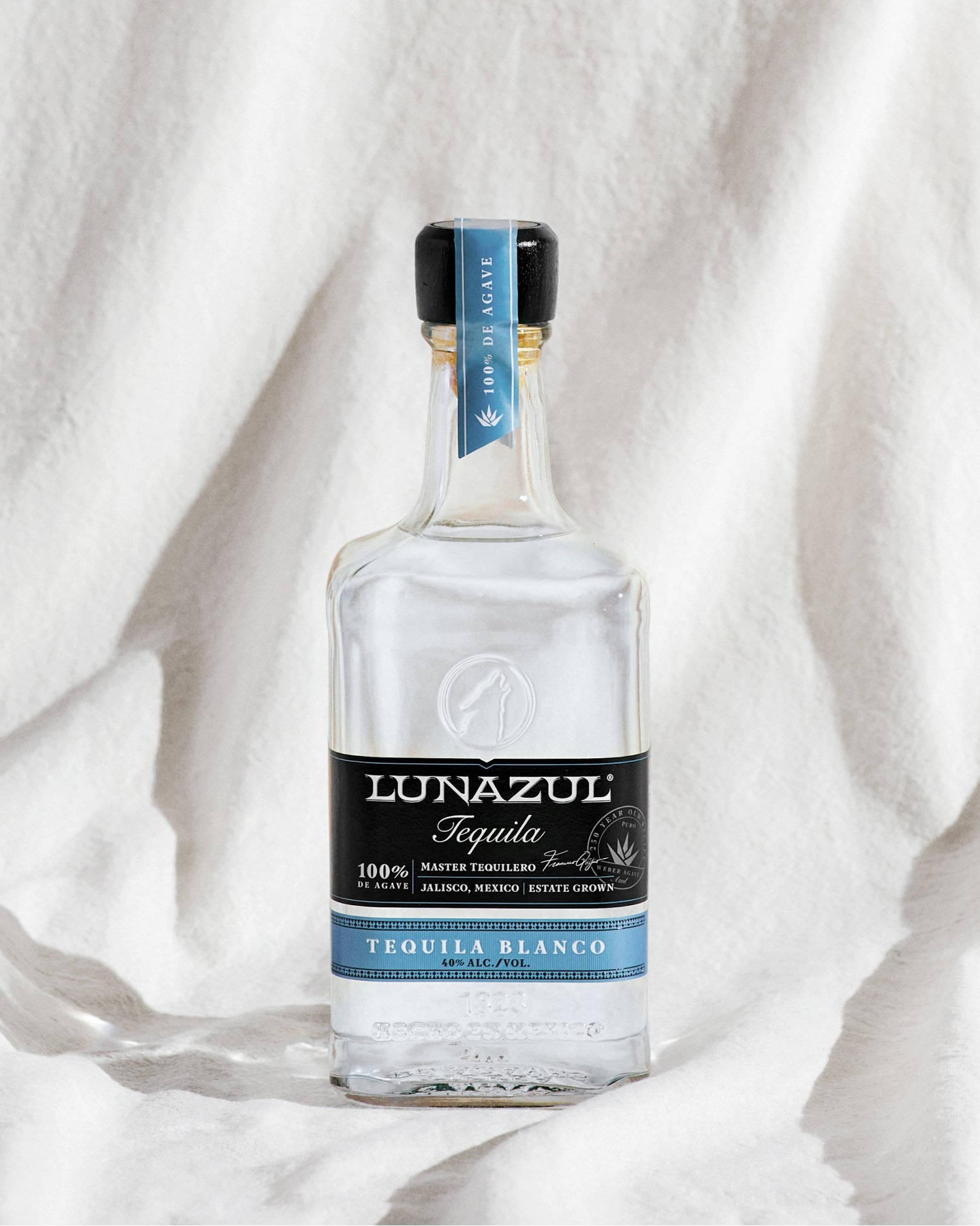 alcohol 桌布 壁紙 — 一支透明的Lunazul Tequila Blanco酒瓶放在柔軟的白色布料上，在明亮的自然光下。