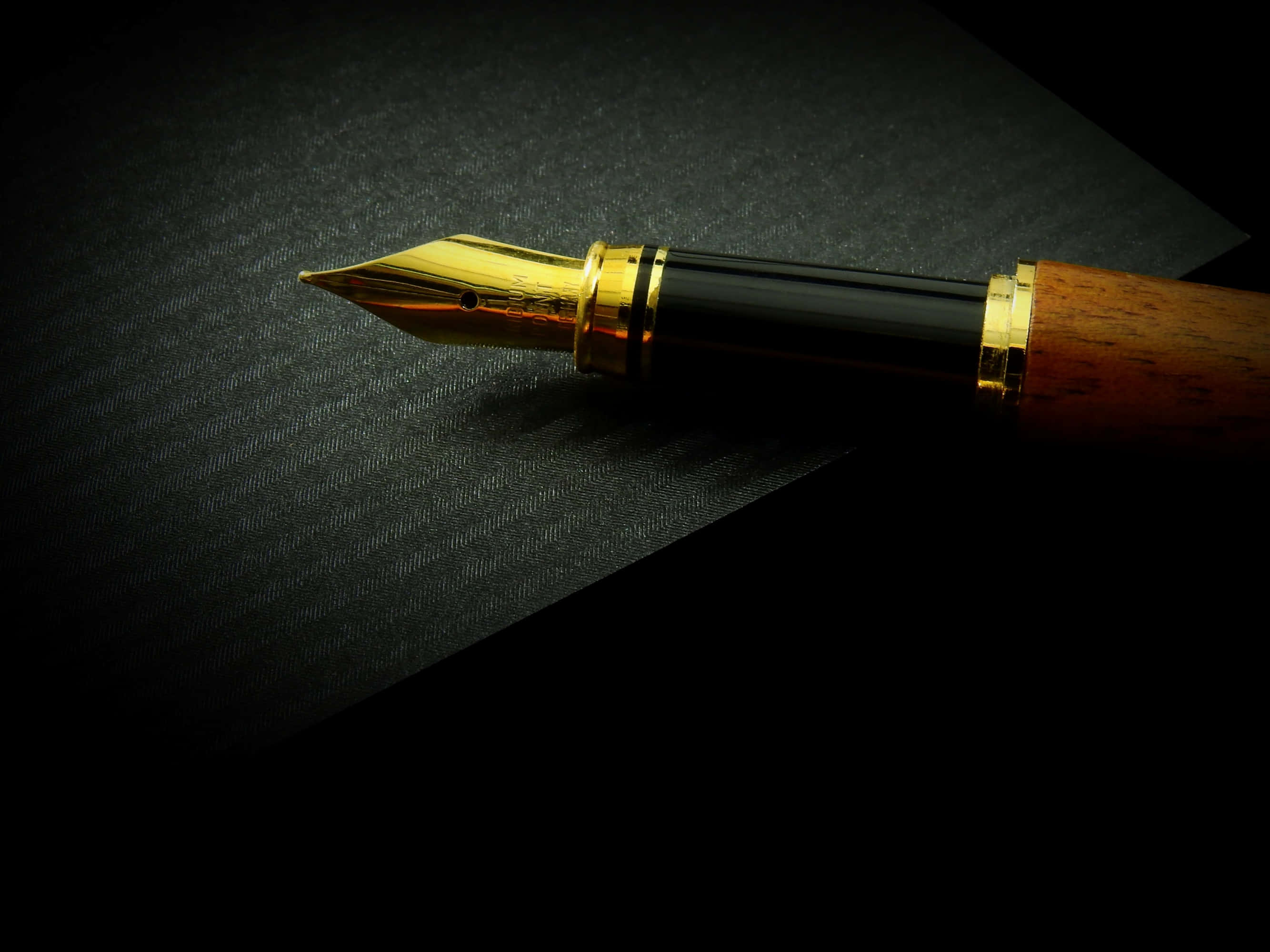 Pen 桌布 壁紙 — 一支金色鋼筆搭配木質筆握，在戲劇性聚光燈下斜放在有紋理的黑色紙上。