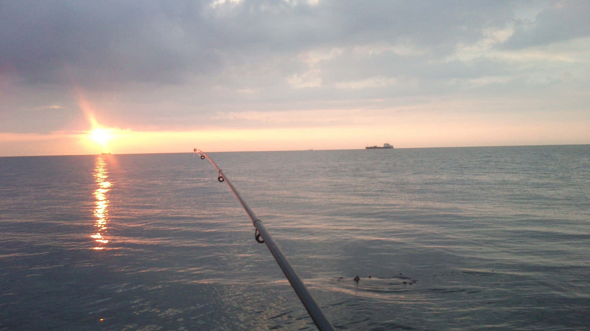 Fishing Rod 桌布 壁紙 — 一支釣竿伸向平靜的海面，太陽在雲層後落下，投射出金色的倒影。