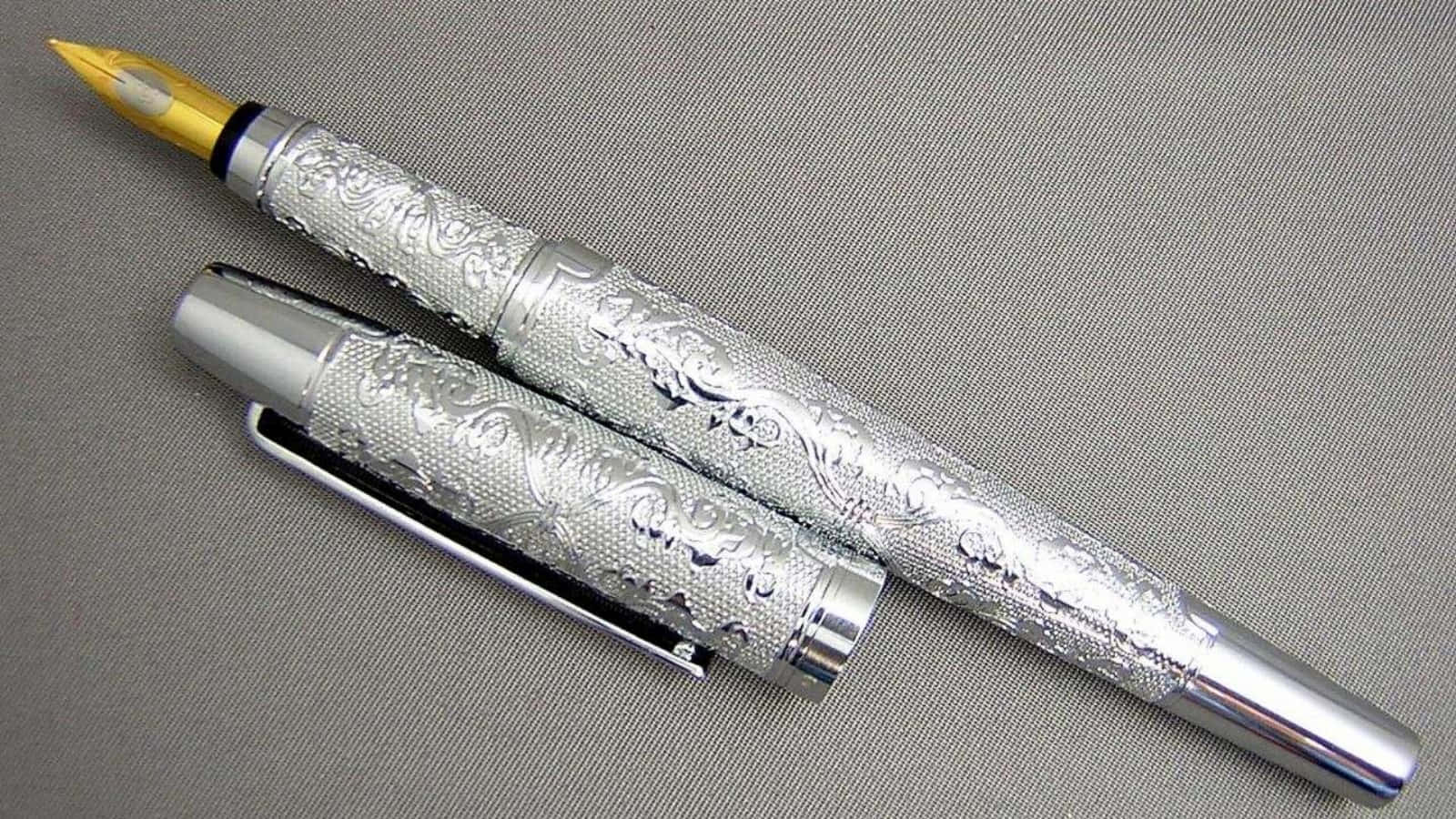 Pen 桌布 壁紙 — 一支銀色鋼筆，帶有精緻的花卉雕刻和金色筆尖，放在有紋理的灰色布料上，旁邊是筆蓋。