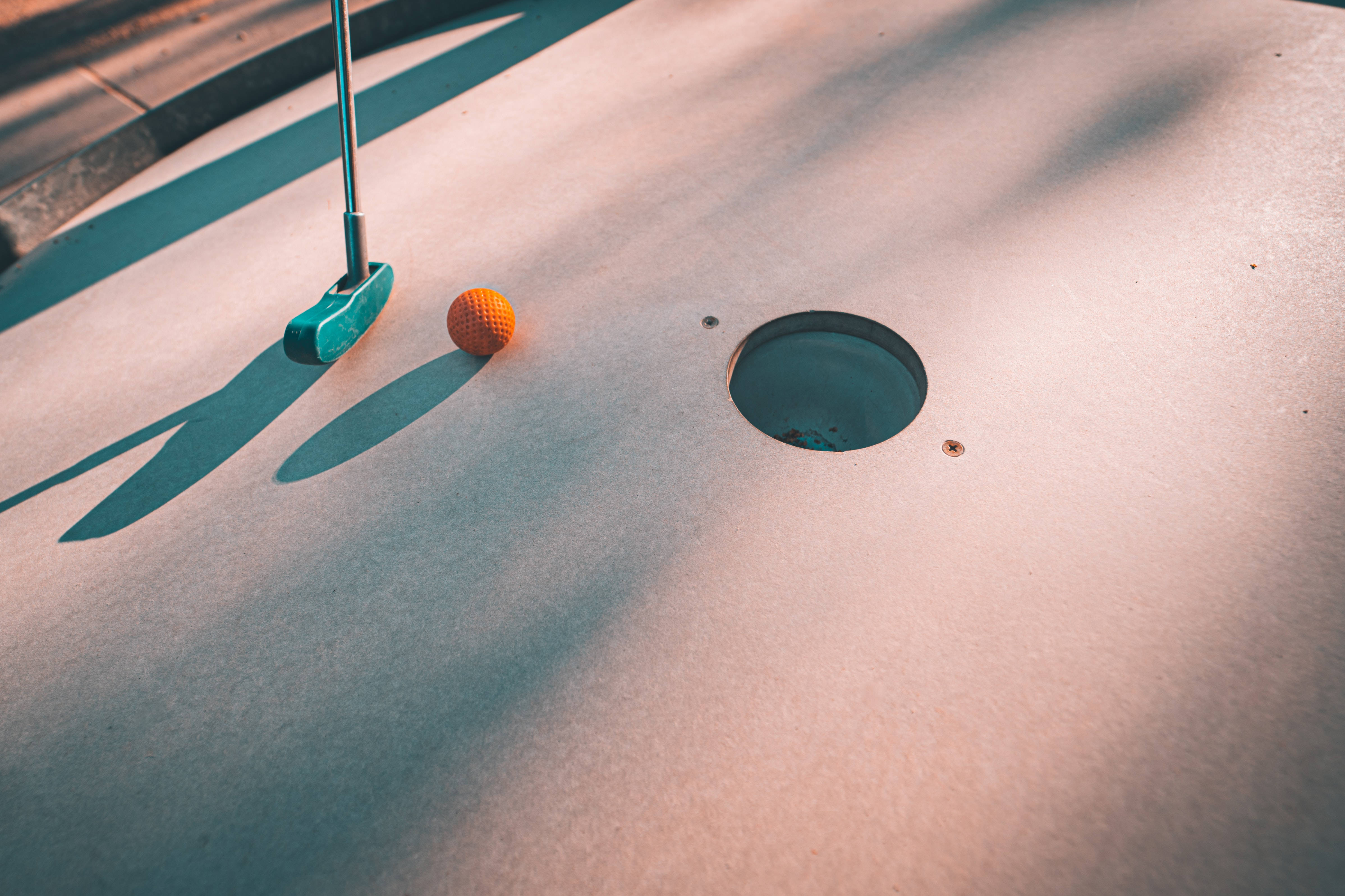 4K Golf 桌布 壁紙 — 一支青綠色推桿和一個橙色高爾夫球放在陽光普照的小型高爾夫球場洞口附近，帶有長長的影子。