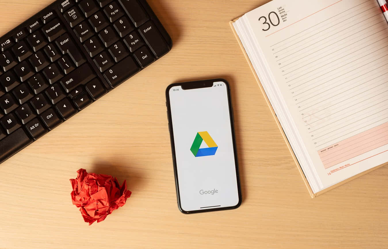 美學 桌布 壁紙 — 一支顯示 Google Drive 的智慧型手機放在木桌上，旁邊有鍵盤、皺巴巴的紅紙和一本打開的計劃簿。