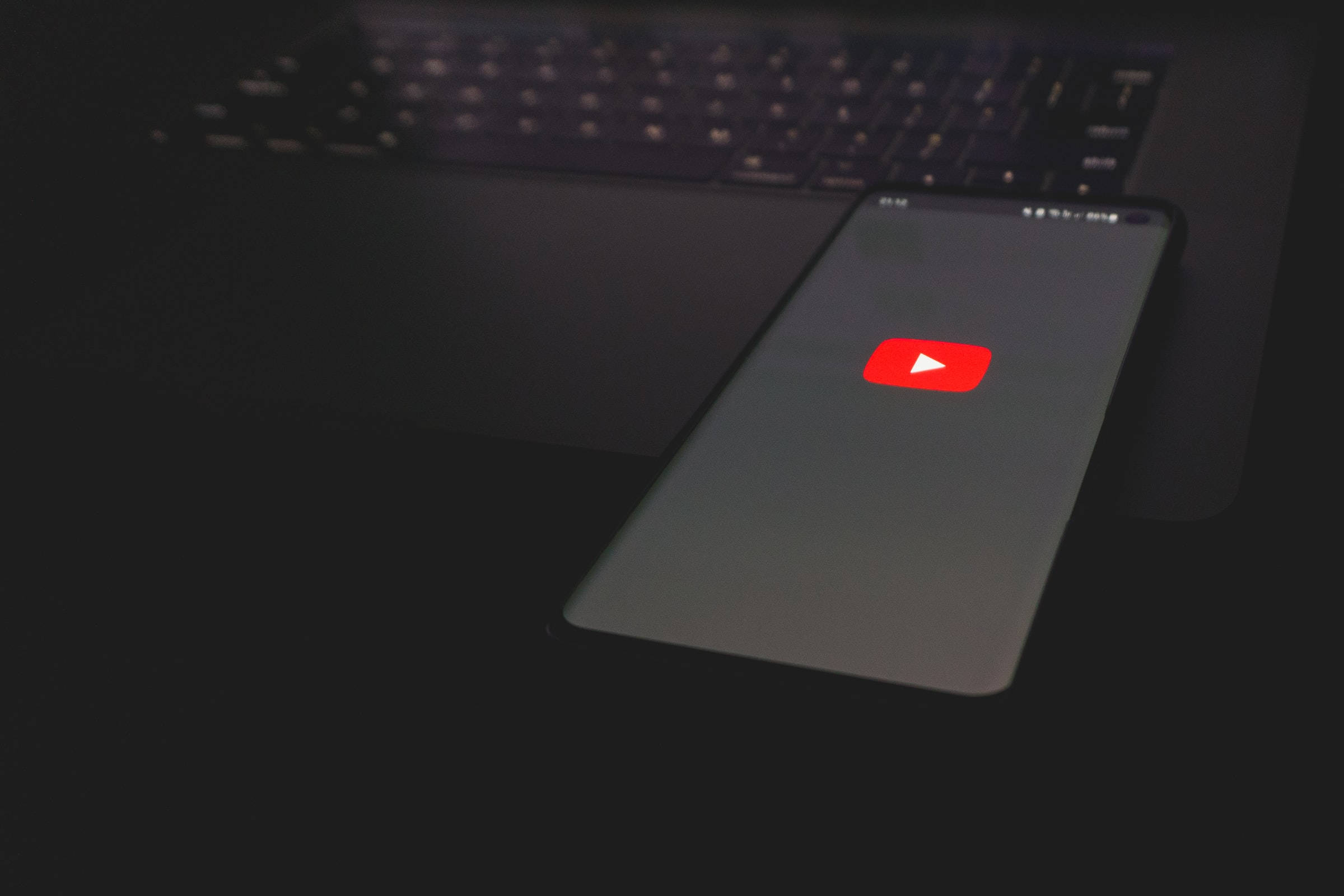Application 桌布 壁紙 — 一支顯示 YouTube 標誌的智慧型手機放在昏暗環境中的深色筆記型電腦鍵盤上。