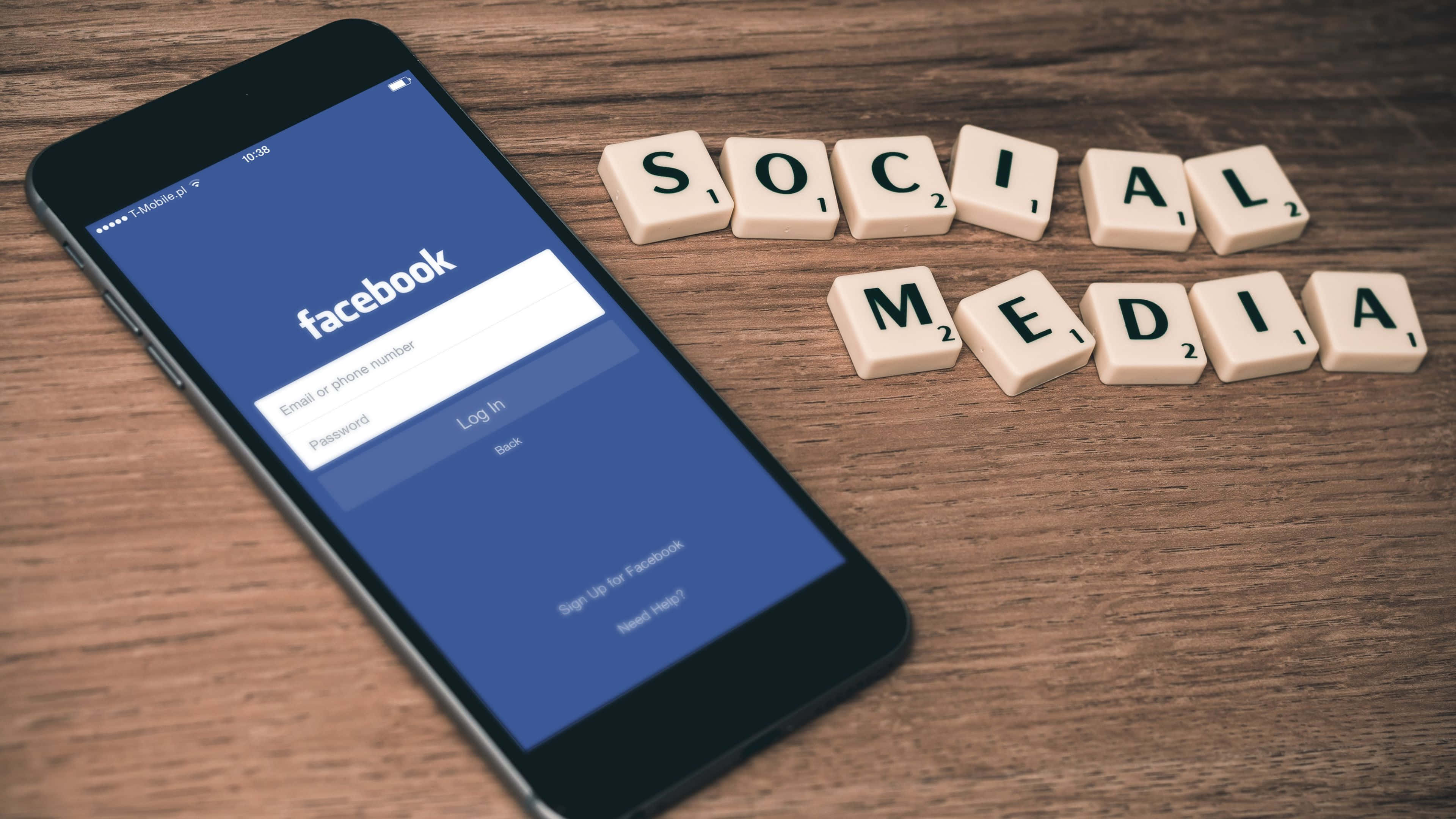 Facebook 桌布 壁紙 — 一支顯示Facebook登入畫面的智慧型手機，旁邊放著拼出「SOCIAL MEDIA」的Scrabble字母牌，置於木質表面上。