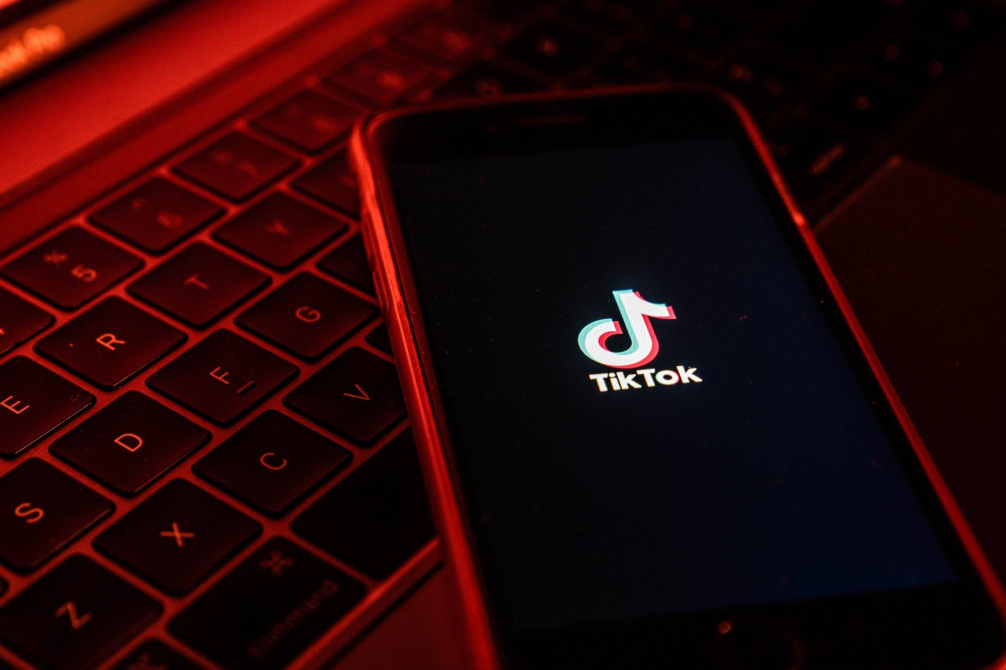 手機 桌布 壁紙 — 一支顯示TikTok標誌的智慧型手機放在一個浸浴在深紅色環境光的筆記型電腦鍵盤上。