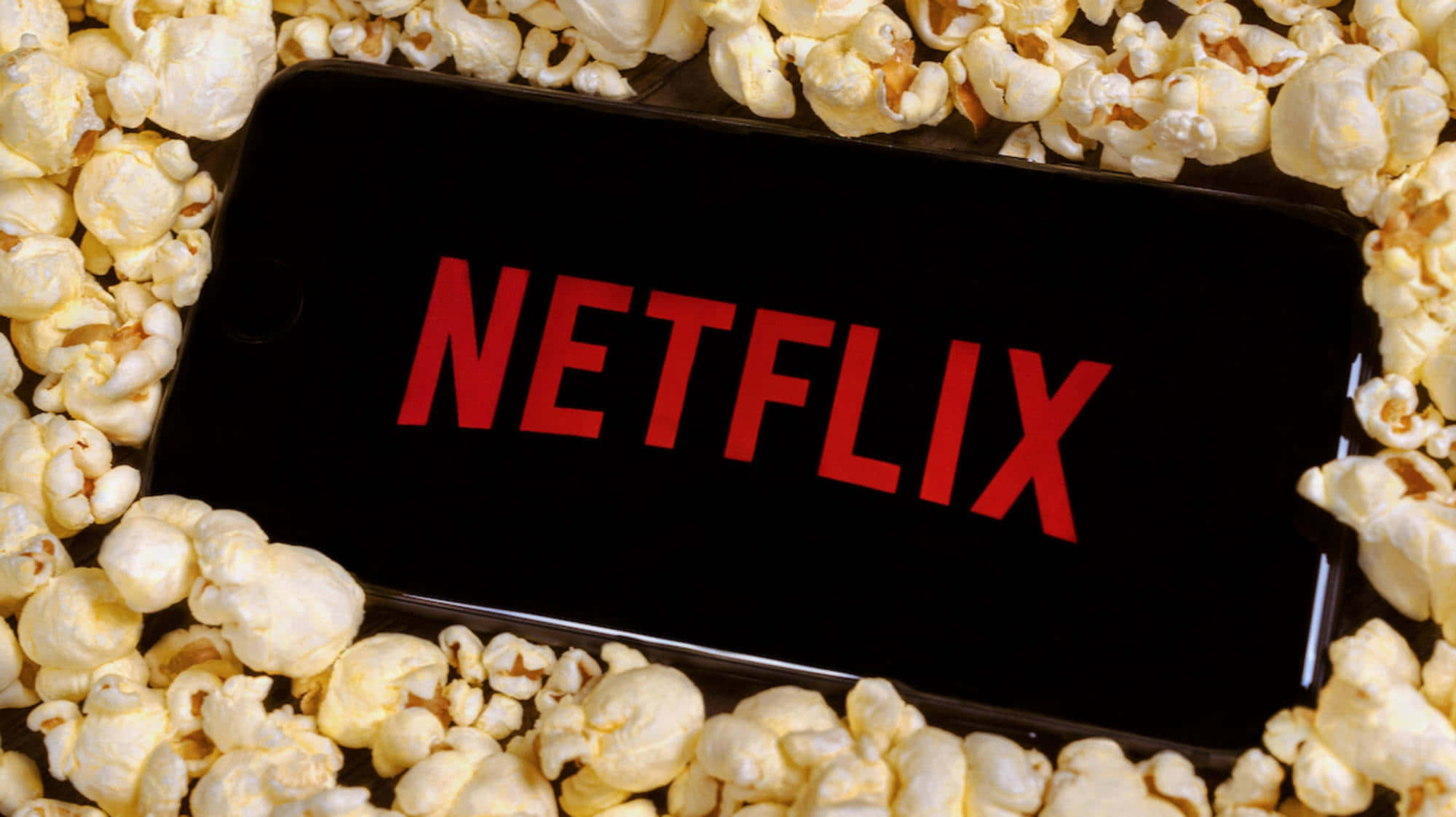 手機 桌布 壁紙 — 一支顯示紅色 Netflix 標誌的智慧型手機，被一層蓬鬆的白色爆米花包圍。