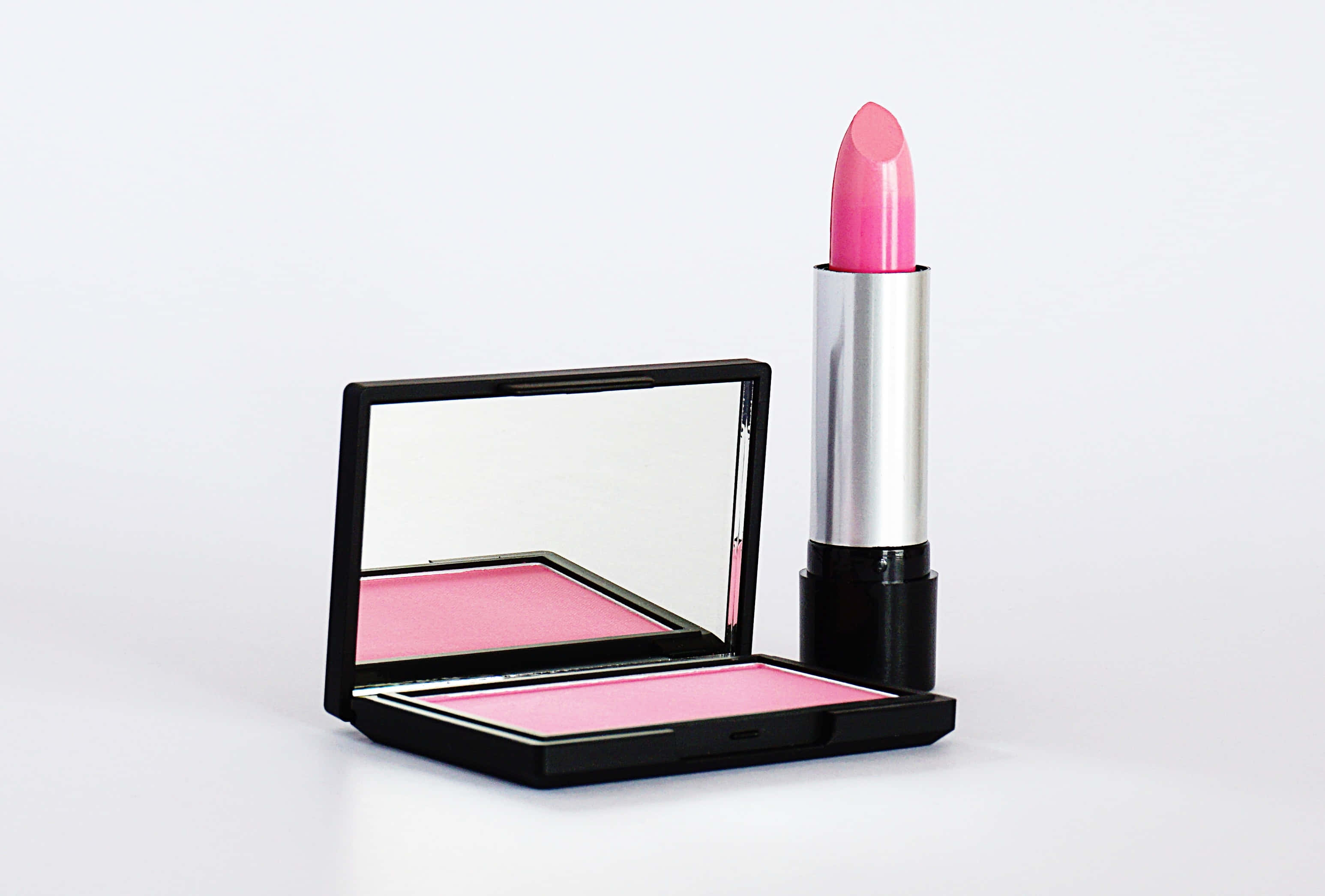 Pink Lipstick 桌布 壁紙 — 一支鮮粉紅色唇膏放在一個打開的黑色粉盒旁邊，盒內有相配的腮紅和鏡子，置於潔白表面上。