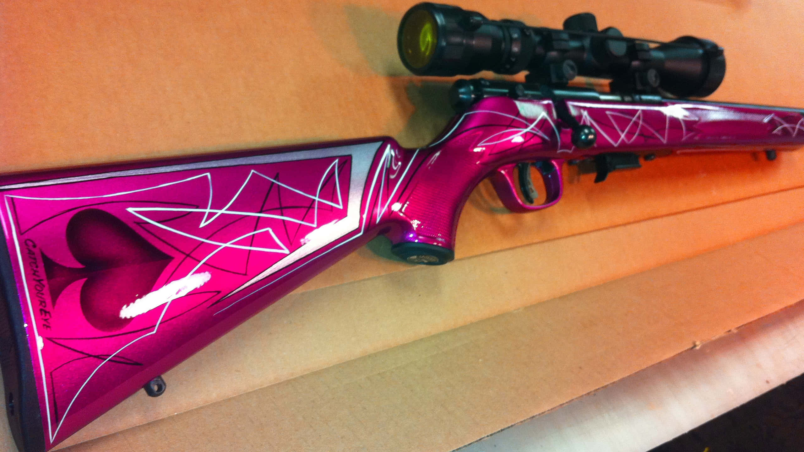 Rifle 桌布 壁紙 — 一支鮮豔的粉紅色定制步槍，帶有白色幾何和心形圖案，放置在木質表面上，在溫暖的燈光下。