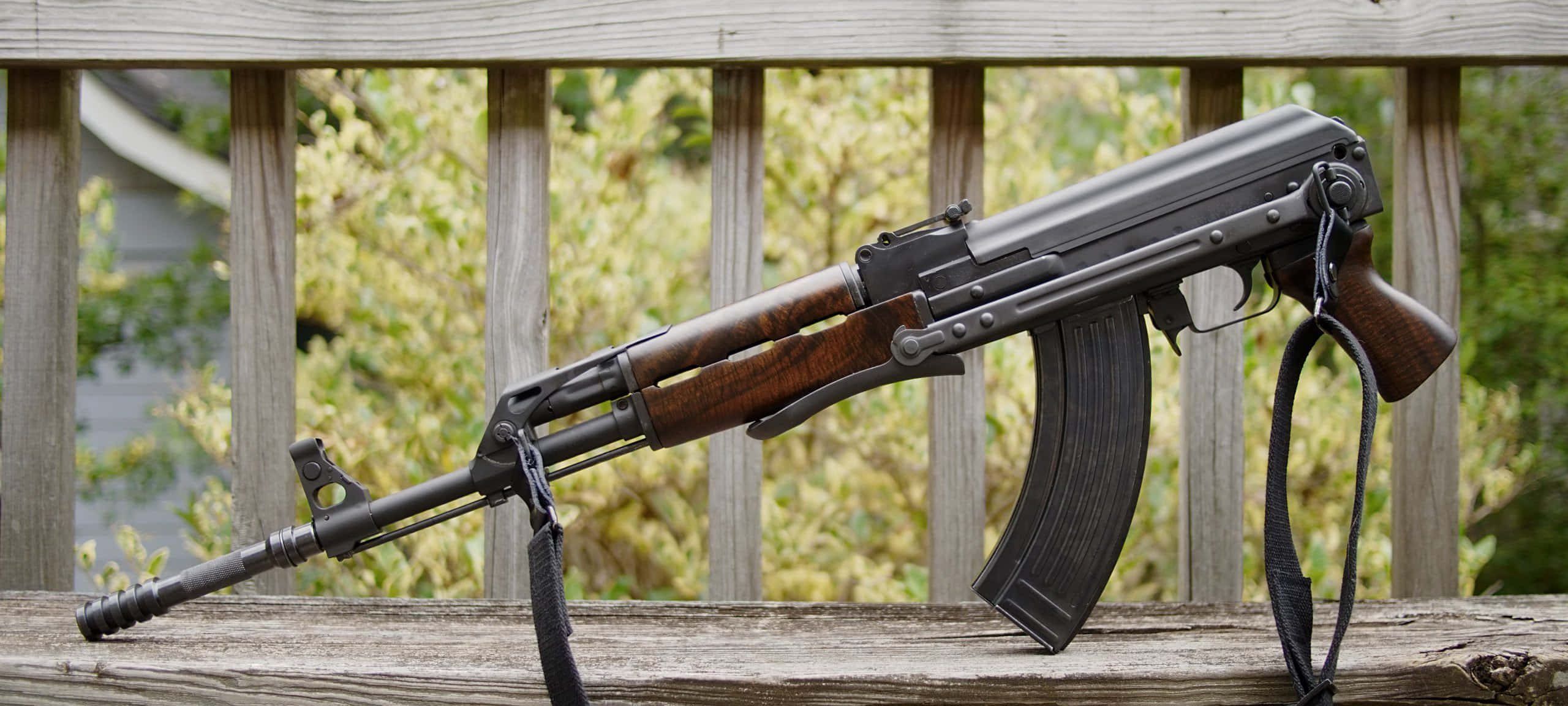 Rifle 桌布 壁紙 — 一支黑色AK-47步槍搭配木質槍托，擱在風化的木製甲板欄杆上，背景是模糊的綠色樹葉。