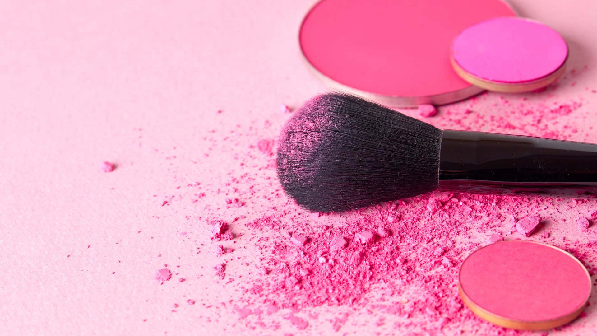 Pink Makeup 桌布 壁紙 — 一支黑色化妝刷躺在柔軟的粉紅色表面上，周圍散落著鮮豔的粉紅色粉末和打開的粉餅盒。