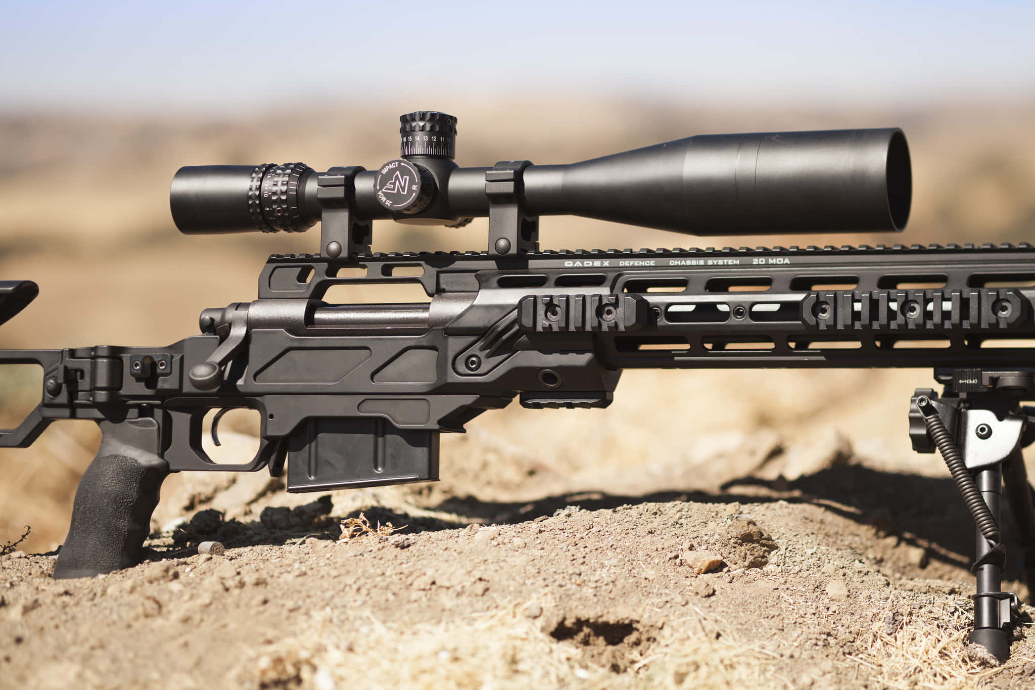 Rifle 桌布 壁紙 — 一支黑色戰術步槍配有瞄準鏡和兩腳架，在明亮的日光下放置在乾燥的土地上。