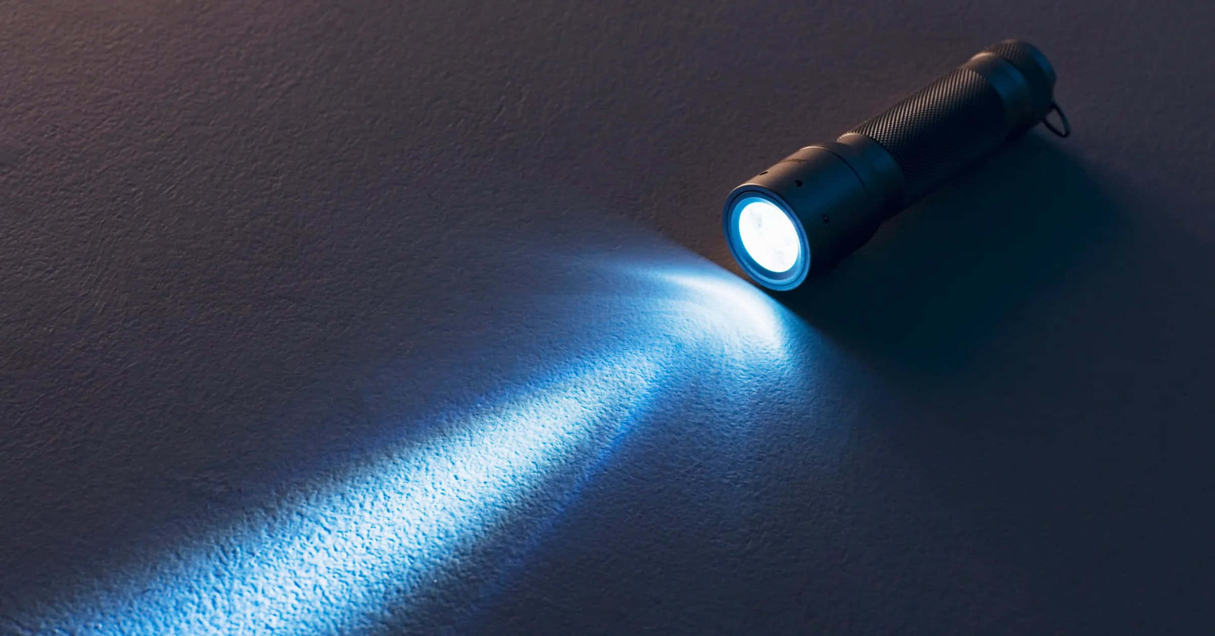 Flashlight 桌布 壁紙 — 一支黑色手電筒在昏暗的環境中，將明亮的藍色光束投射在帶有紋理的深色表面上。