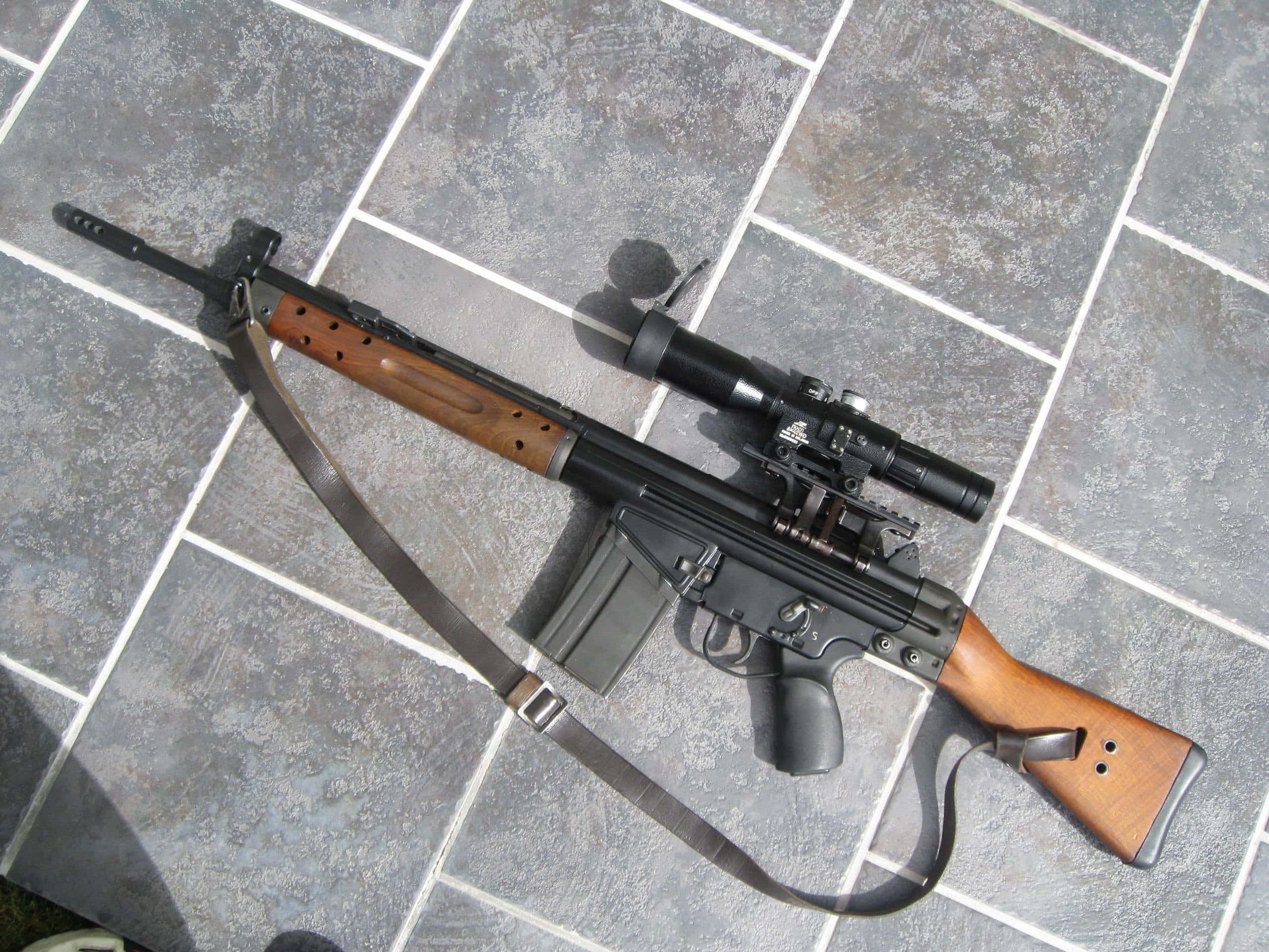 Rifle 桌布 壁紙 — 一支黑色木製步槍配有瞄準鏡和槍帶，在明亮的日光下放在灰色瓷磚地板上。