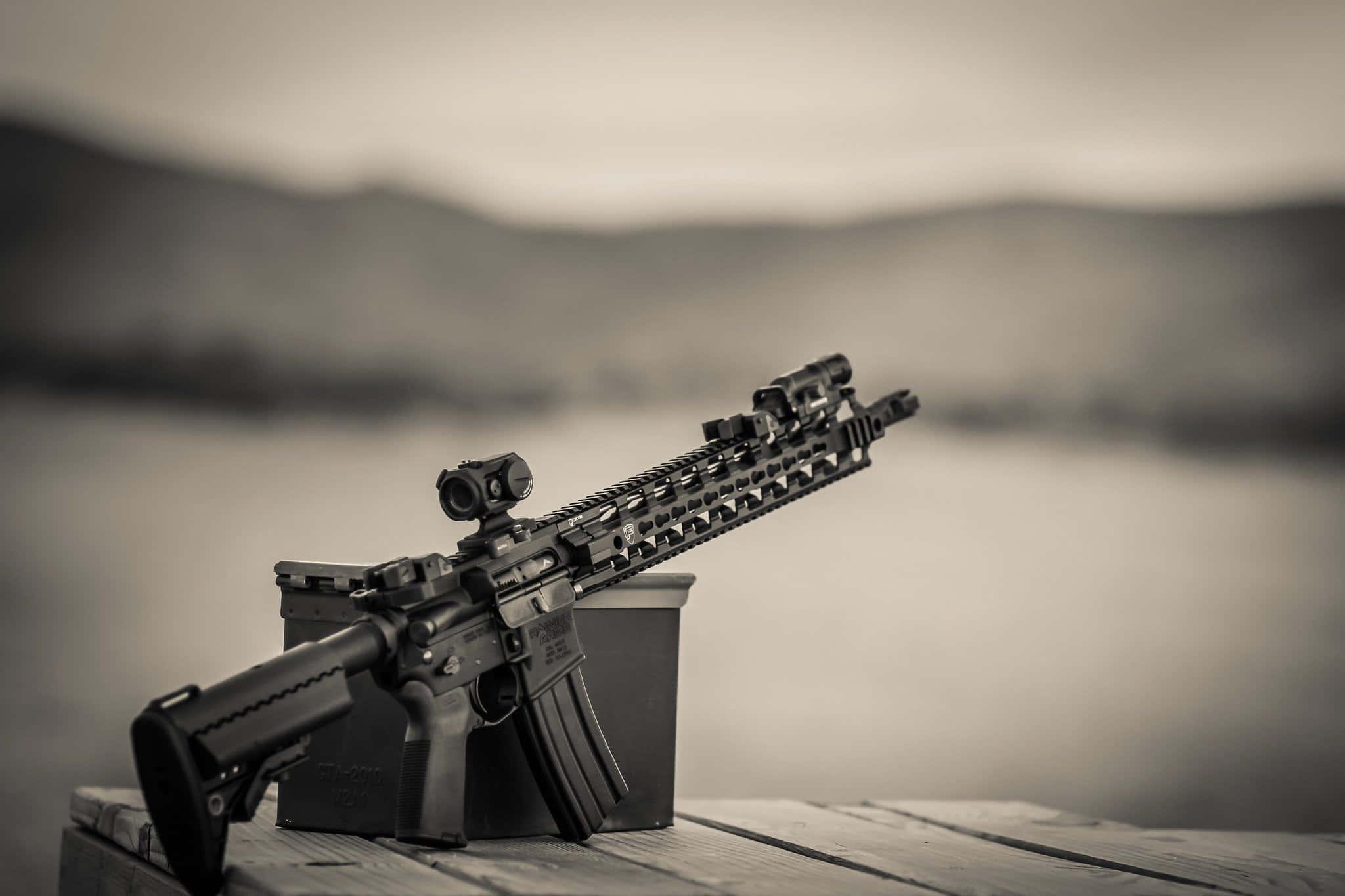 Rifle 桌布 壁紙 — 一支黑色步槍擱在木製碼頭上，背景是模糊的湖泊和山脈，以單色呈現。