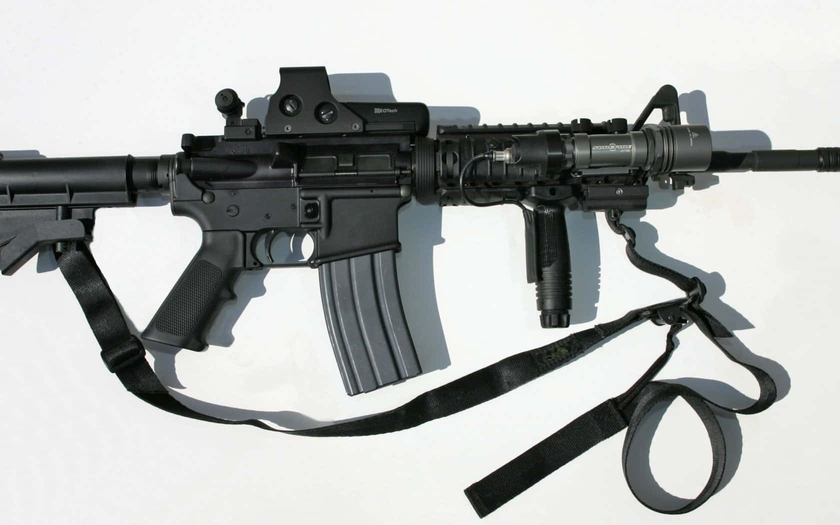Rifle 桌布 壁紙 — 一支黑色突擊步槍配有戰術配件，放置在純白表面上，投射出銳利的陰影。