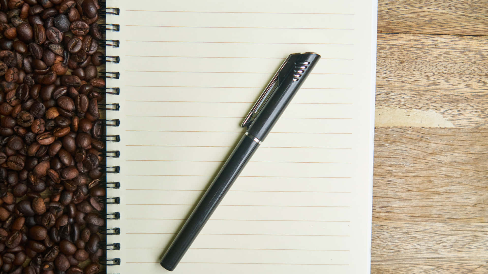 notes 桌布 壁紙 — 一支黑色筆斜放在一本有橫線的筆記本上，旁邊是一堆烘焙咖啡豆在木質表面上。