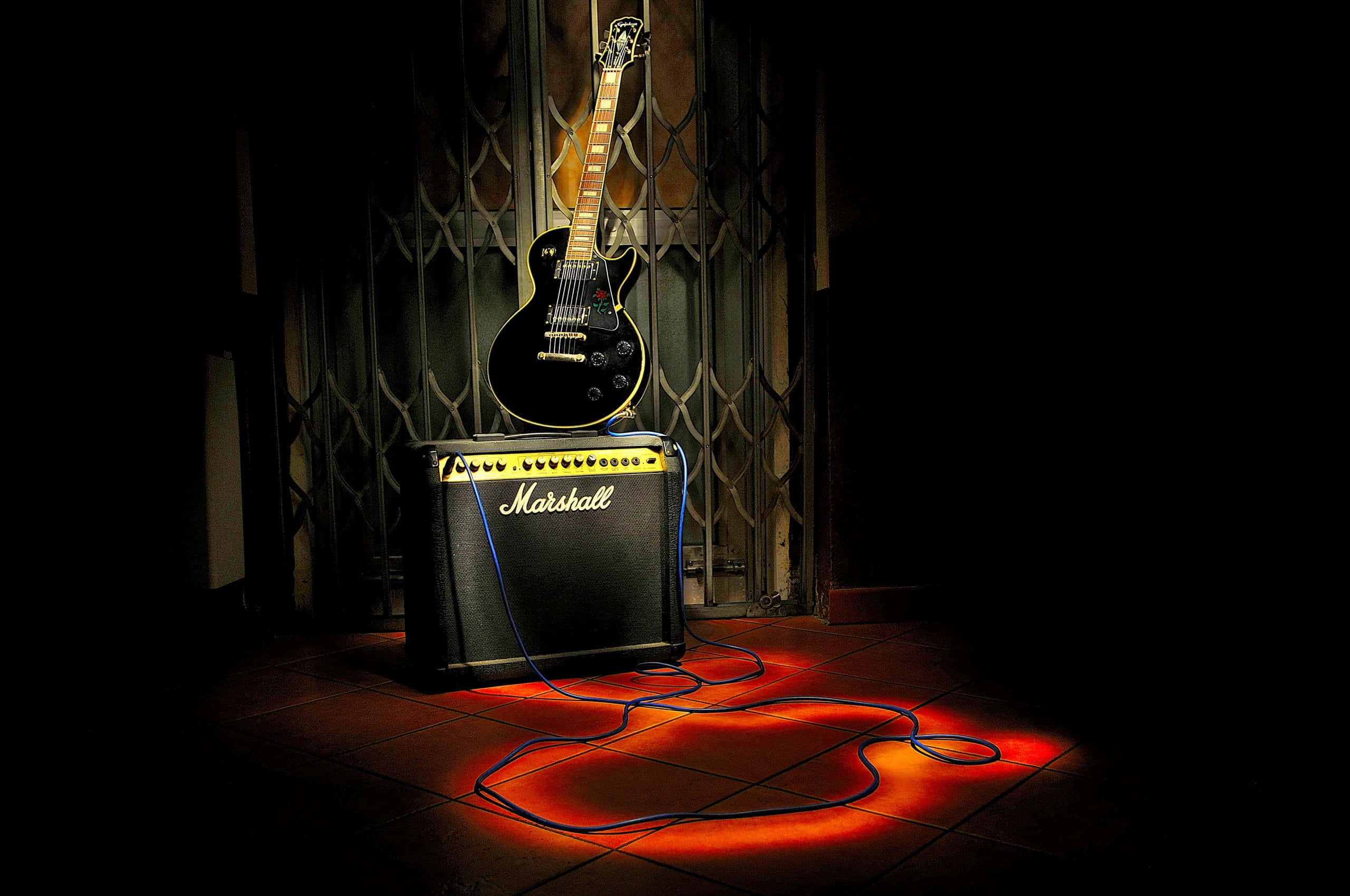Marshall 桌布 壁紙 — 一支黑色電吉他靠在金屬閘門上，閘門上方有一個Marshall擴音機，在昏暗光線下於磁磚地板上投射出溫暖的橙色光芒。