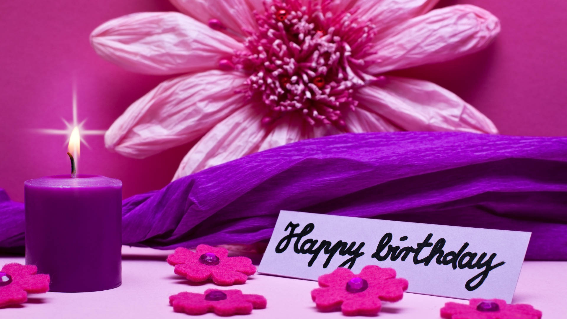 Happy Birthday Flower 桌布 壁紙 — 一支點燃的紫色蠟燭，旁邊有一朵大粉紅花和一張被粉紅紙花包圍的「Happy Birthday」賀卡。
