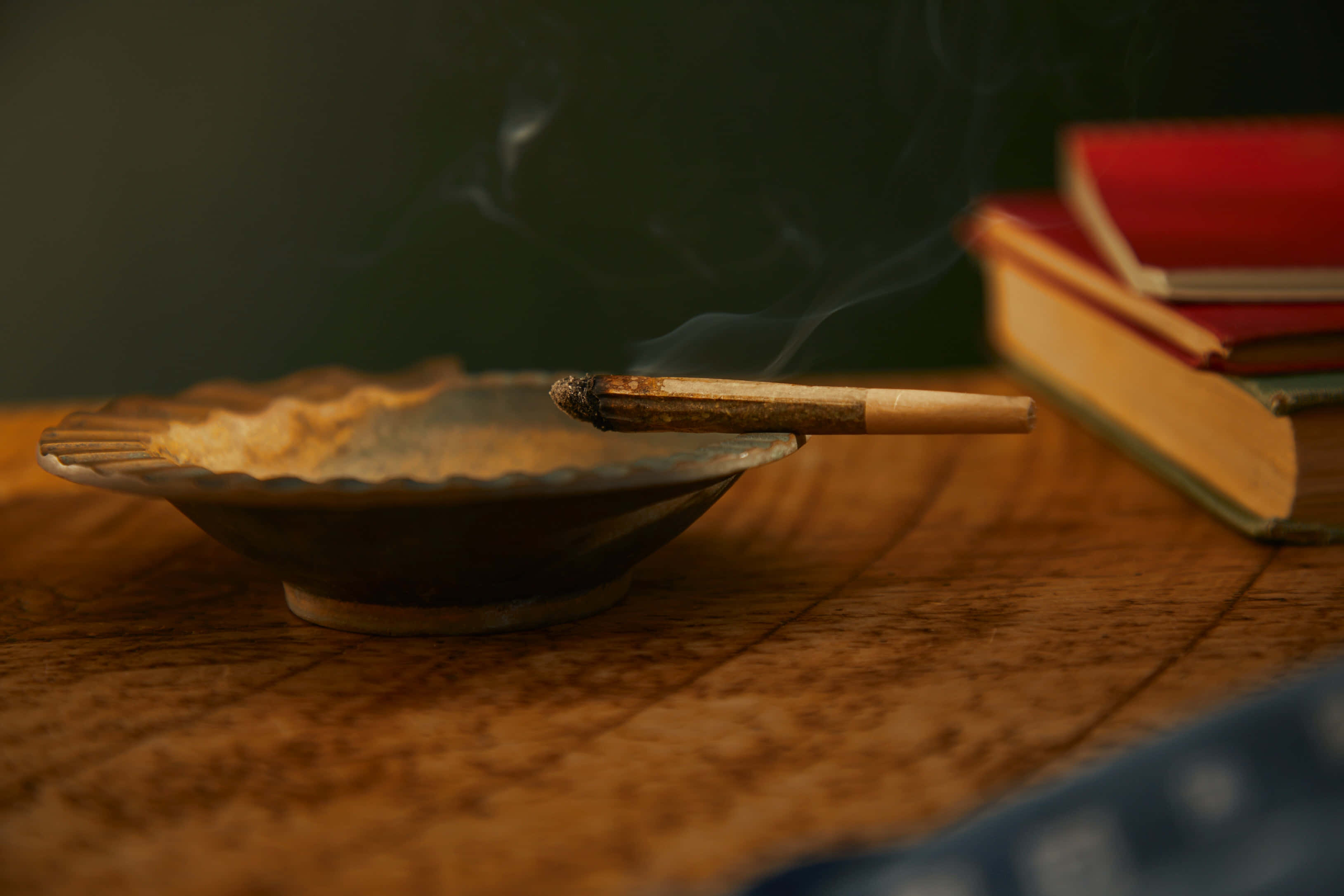 Weed Blunt 桌布 壁紙 — 一支點燃的雪茄放在木桌上的陶瓷煙灰缸裡，旁邊是堆疊的書籍，煙霧在溫暖的環境光中向上繚繞。