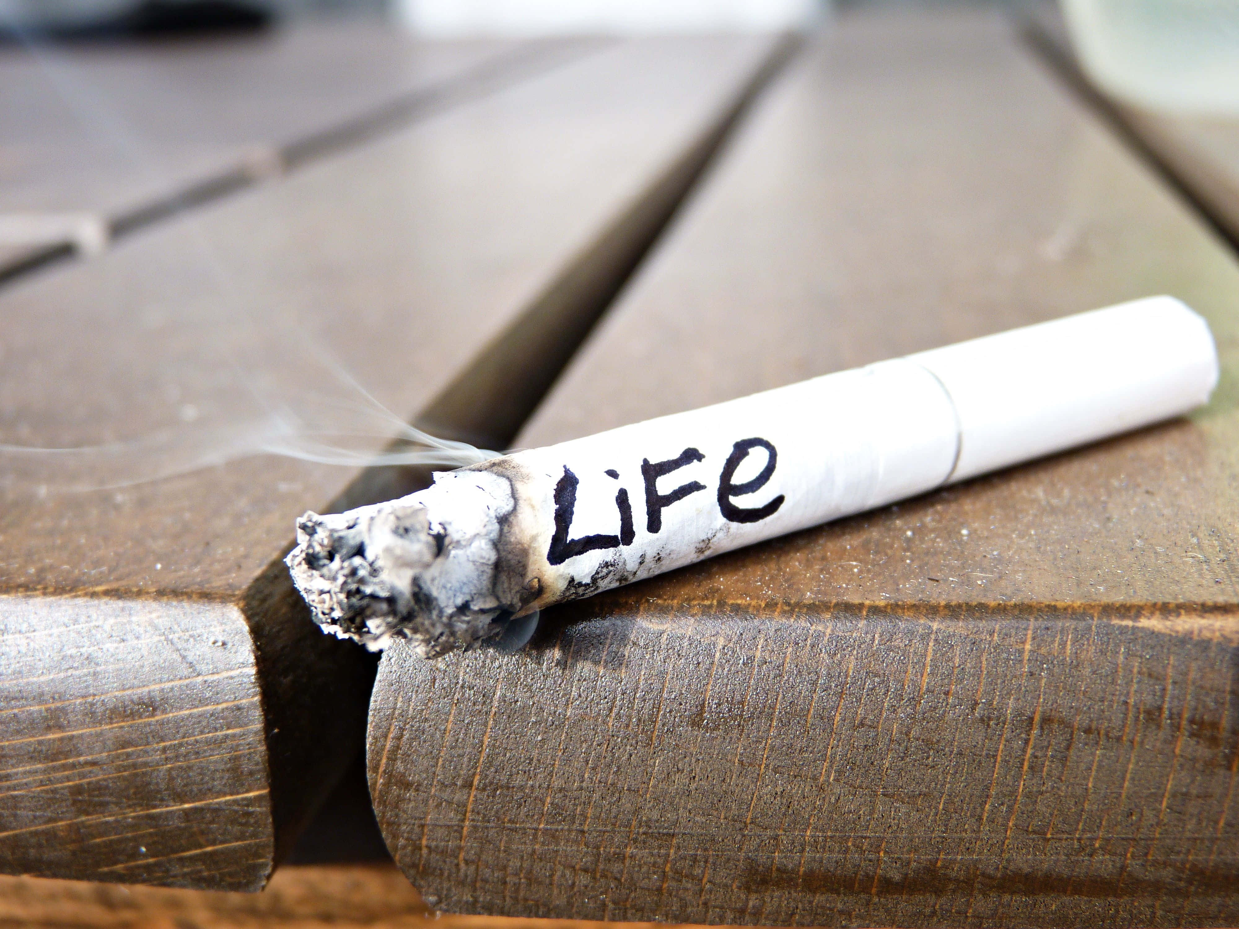 Butt 桌布 壁紙 — 一支點燃的香煙上寫著「Life」，在風化的木質表面上輕輕冒煙。