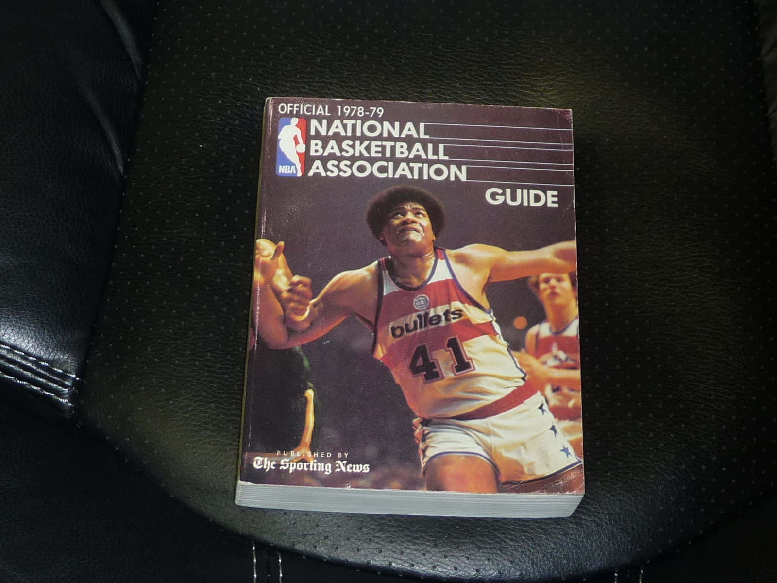 籃球 桌布 壁紙 — 一本1978-79年復古NBA指南，上面有穿著子彈隊球衣的籃球運動員，放在黑色皮革座椅上。