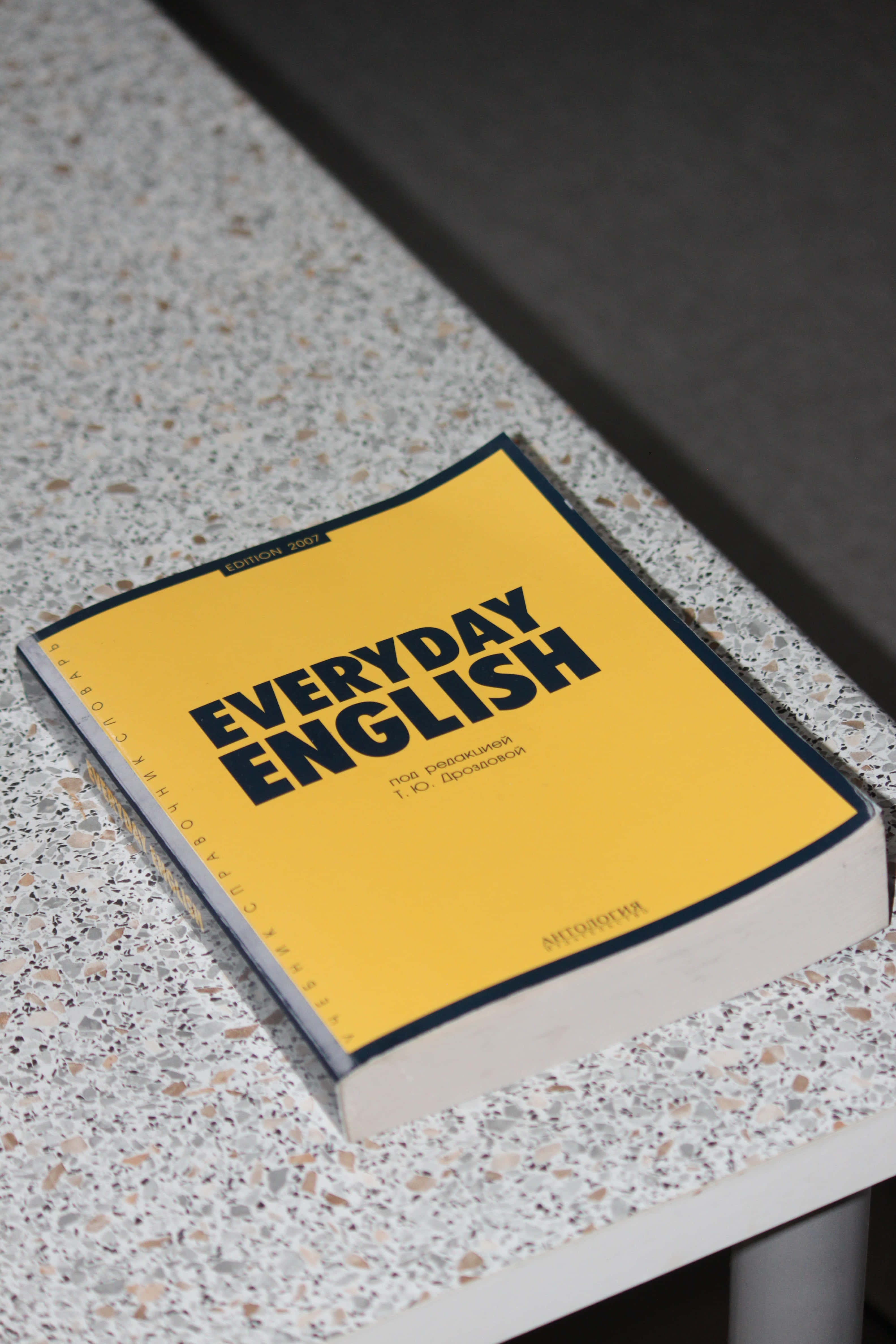 English 桌布 壁紙 — 一本亮黃色的「Everyday English」教科書放在帶有斑點的水磨石表面上，光線柔和自然。