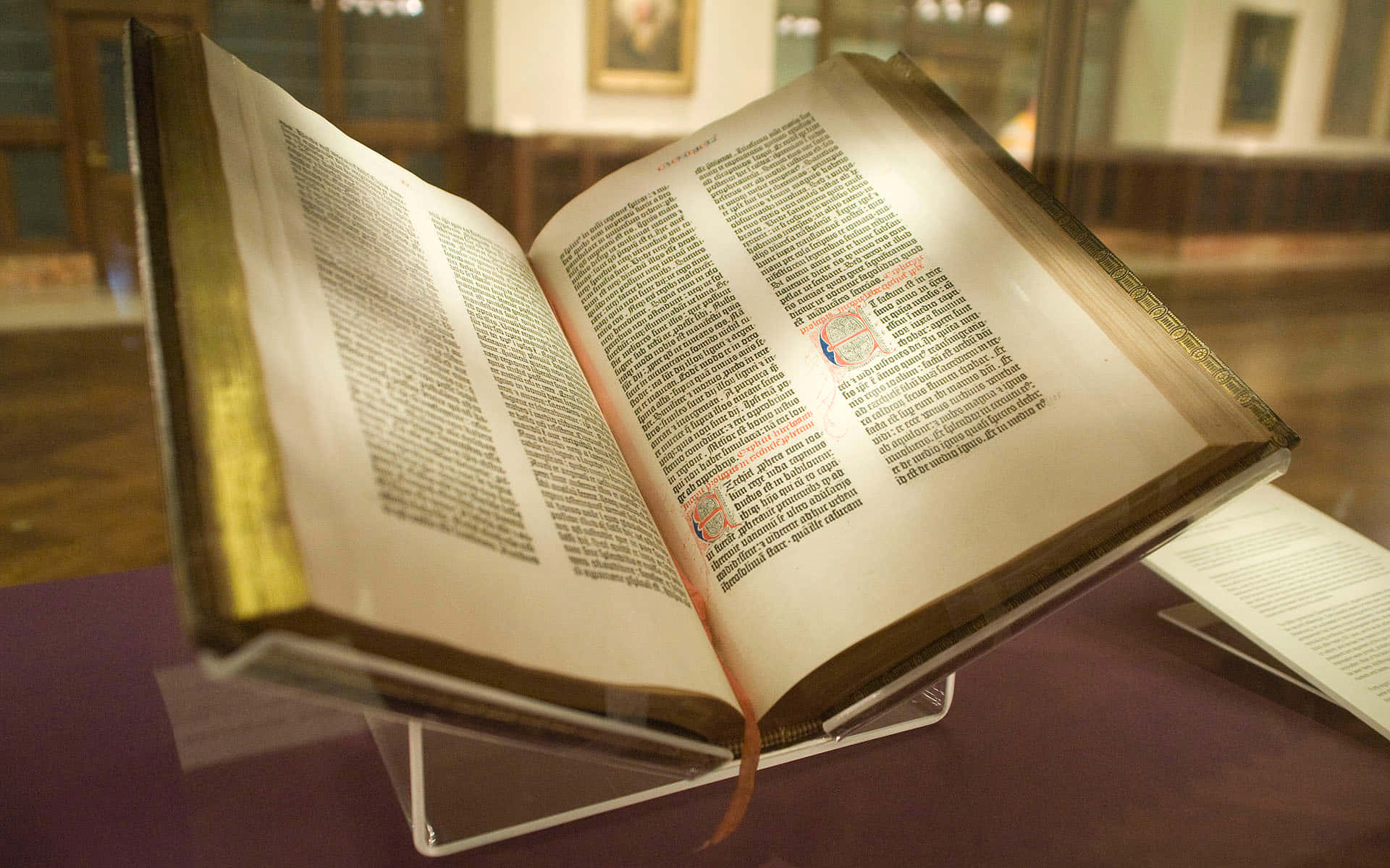 Biblical 桌布 壁紙 — 一本古老的彩繪手稿書，打開展示在玻璃櫃中，有金色邊緣和紅色墨水的首字母。