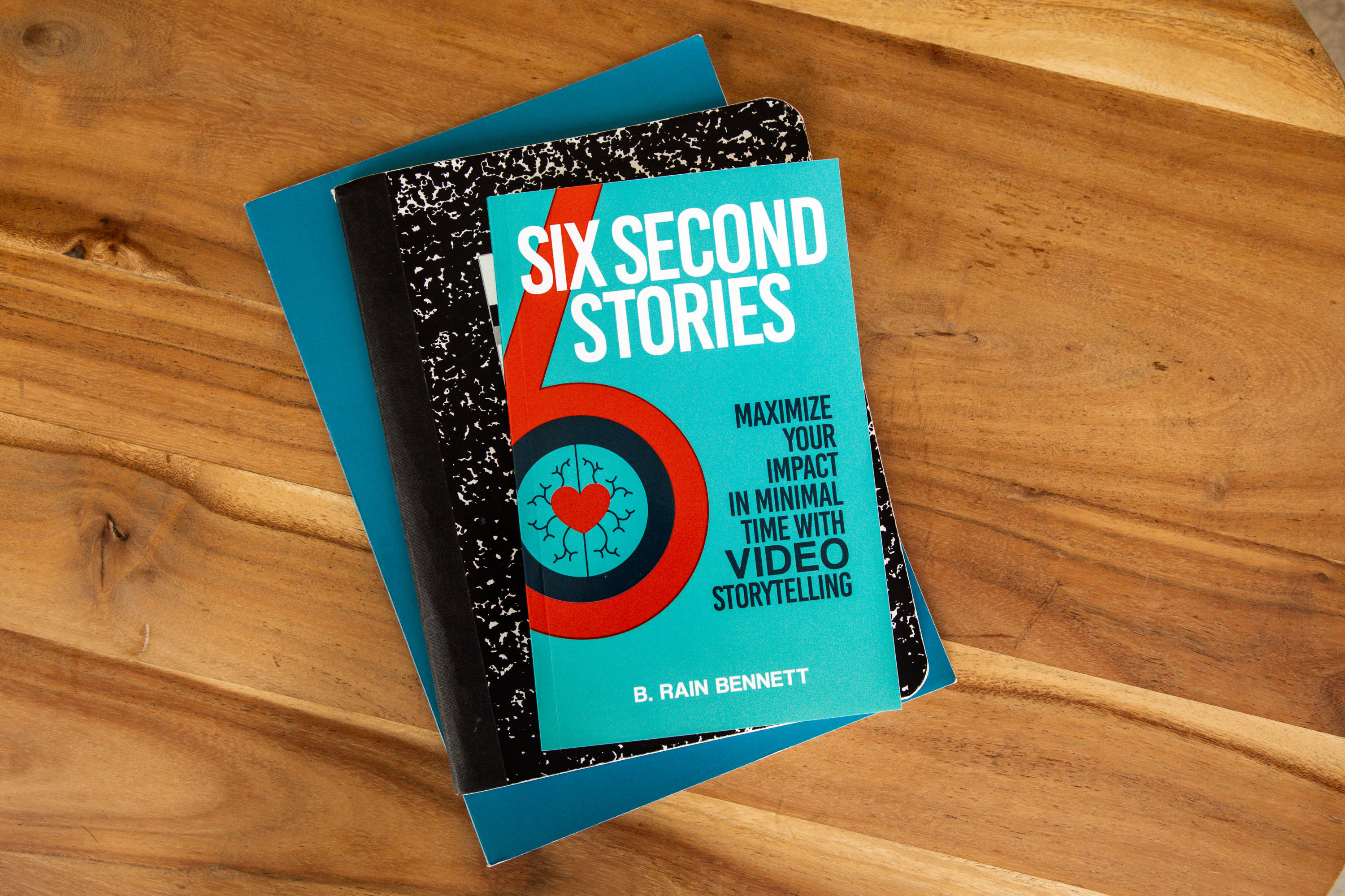 Journals 桌布 壁紙 — 一本名為「Six Second Stories」的藍綠色書本放在藍色筆記本上，置於溫暖的木桌上。