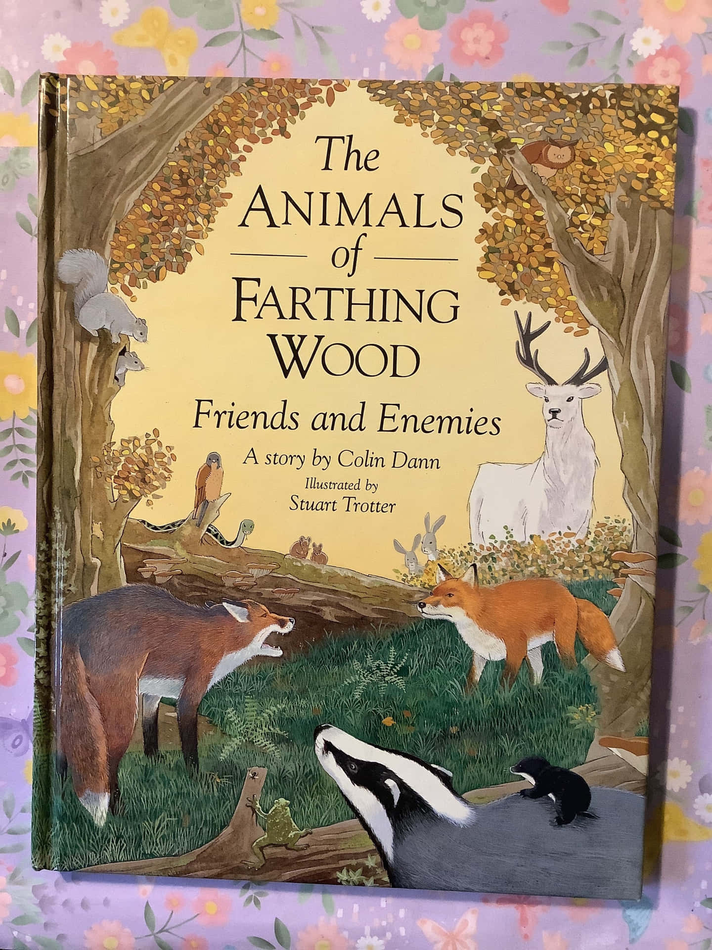 森林 桌布 壁紙 — 一本名為《The Animals of Farthing Wood》的兒童書放在花卉圖案的表面上，展示森林場景中插圖的狐狸、鹿和獾。