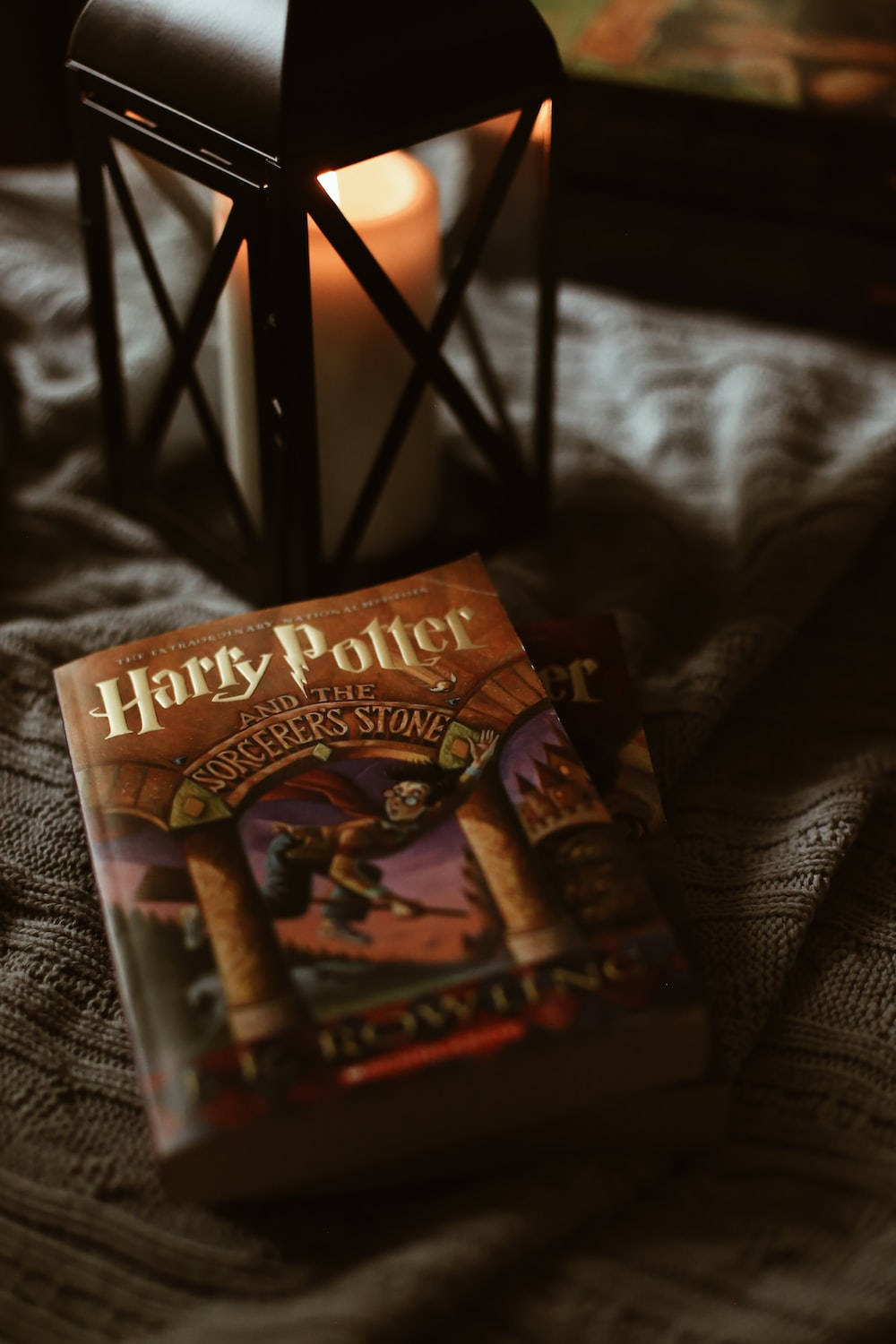 hp aesthetic 桌布 壁紙 — 一本哈利波特書放在有紋理的毯子上，旁邊有一個發光的燈籠，在舒適的室內環境中投射出溫暖的光線。