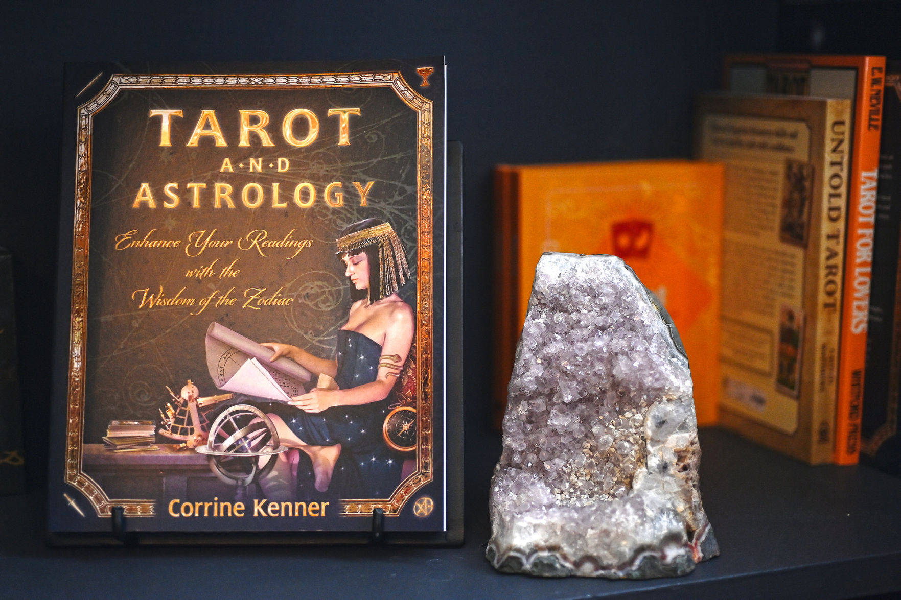 書本 桌布 壁紙 — 一本塔羅牌和占星學書籍放在一個黑暗的架子上，旁邊有一塊閃閃發光的晶洞，還有其他神秘書籍。
