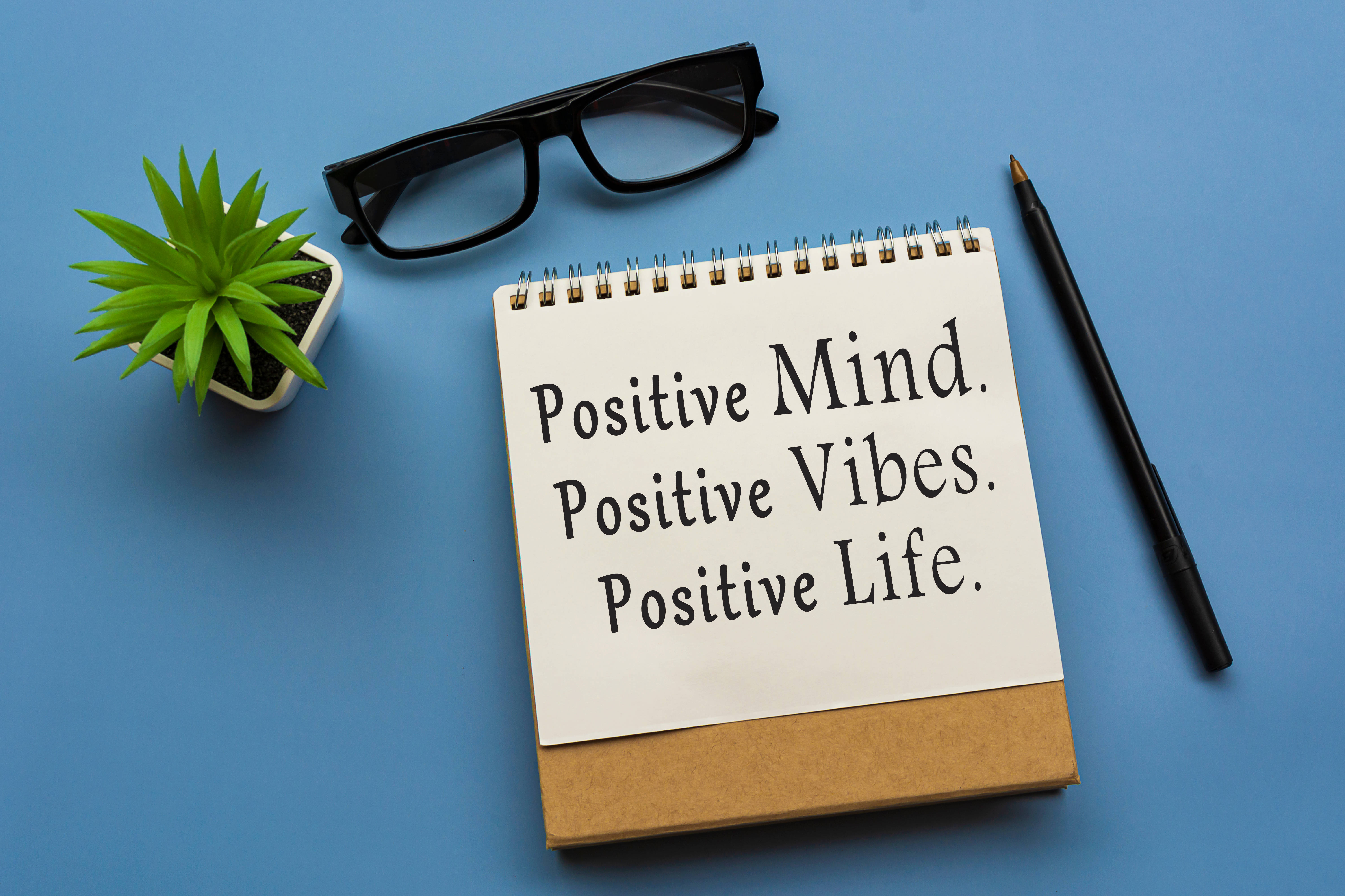 Positive Quotes 桌布 壁紙 — 一本帶有正面肯定語的筆記本放在眼鏡、一支筆和一棵小綠色植物旁邊，置於純藍色表面上。
