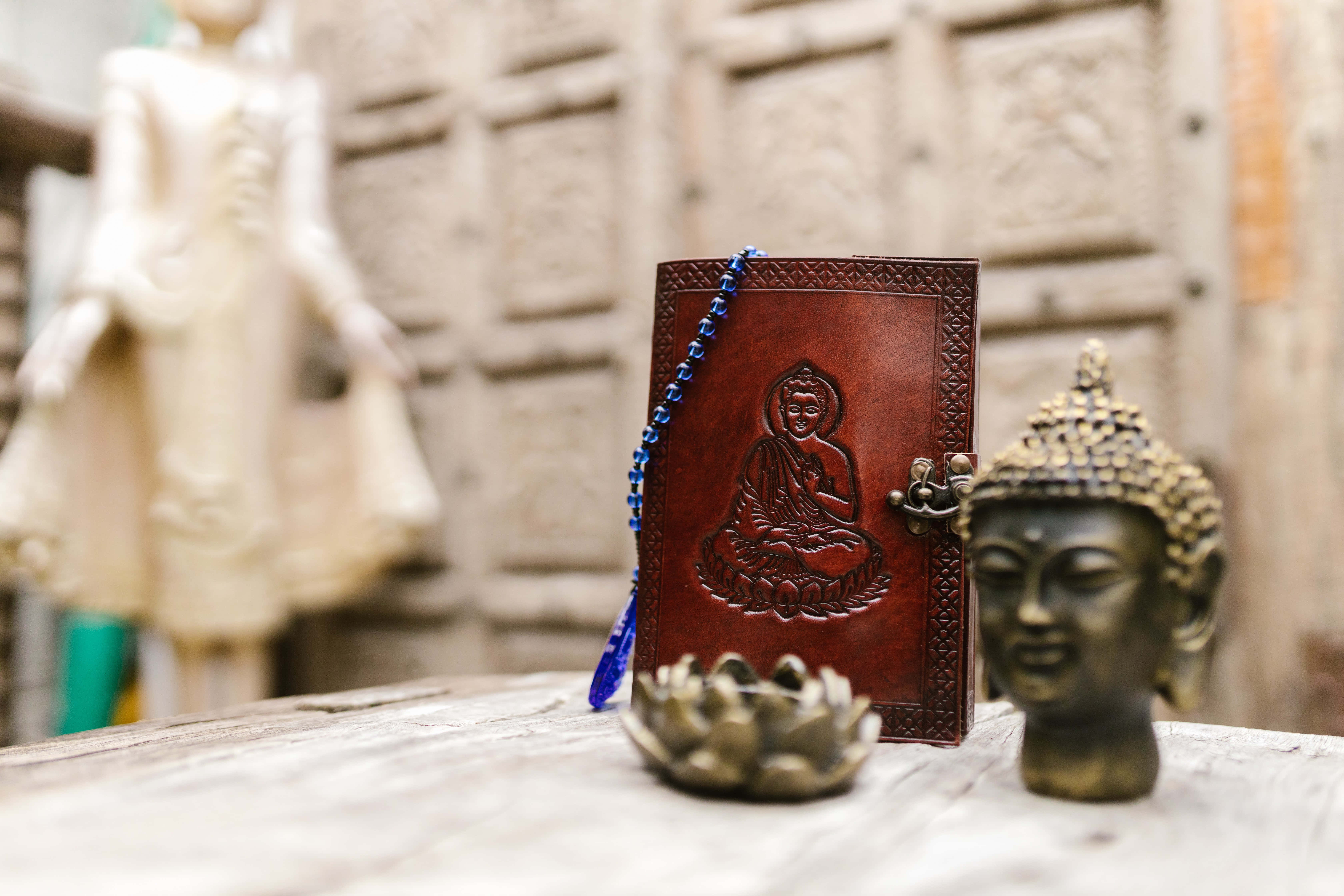 religion 桌布 壁紙 — 一本帶有藍色珠子的皮革裝訂佛陀日記，旁邊放著一個青銅佛陀頭像和蓮花，放在一張風化的木桌上。