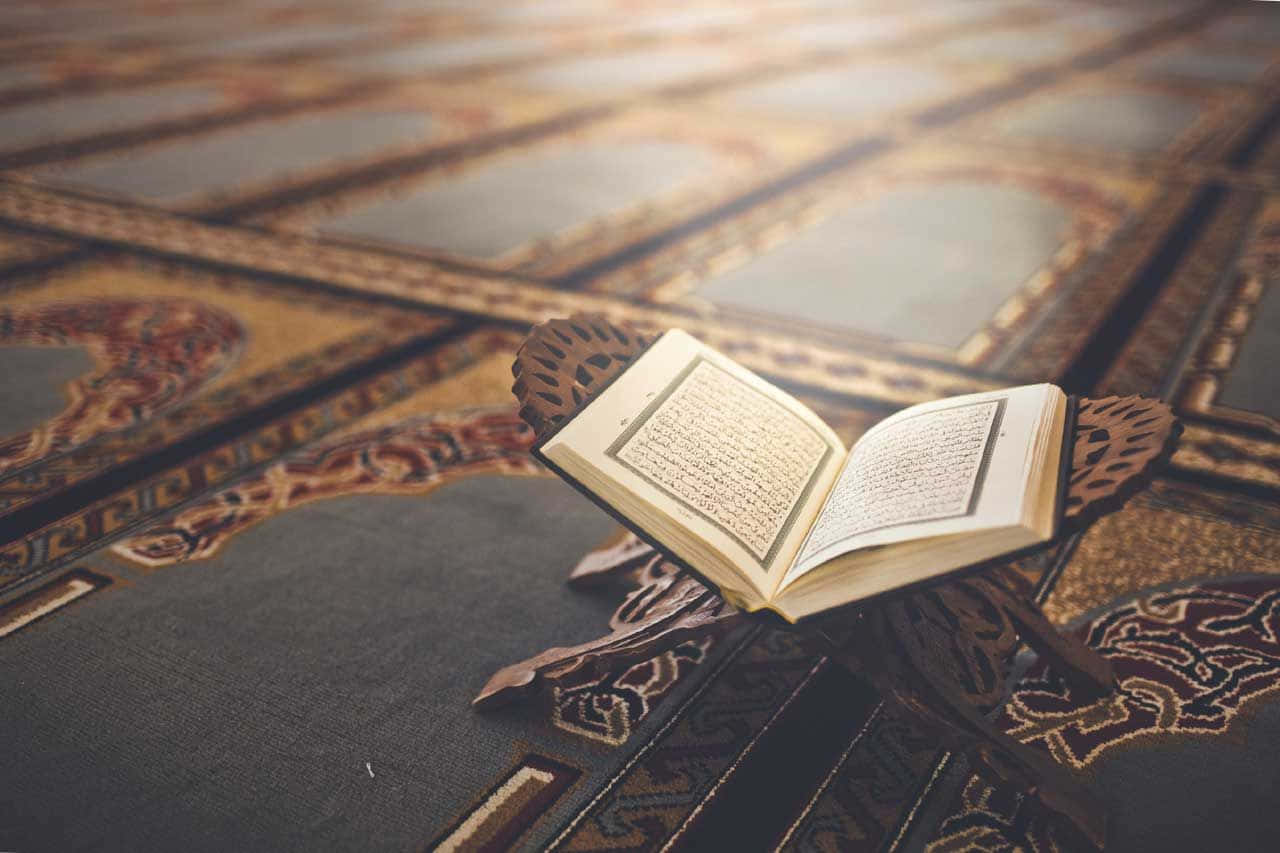 Quran 桌布 壁紙 — 一本打開的古蘭經放在一塊裝飾華麗、帶有精緻金色圖案和深藍色調的祈禱毯上。