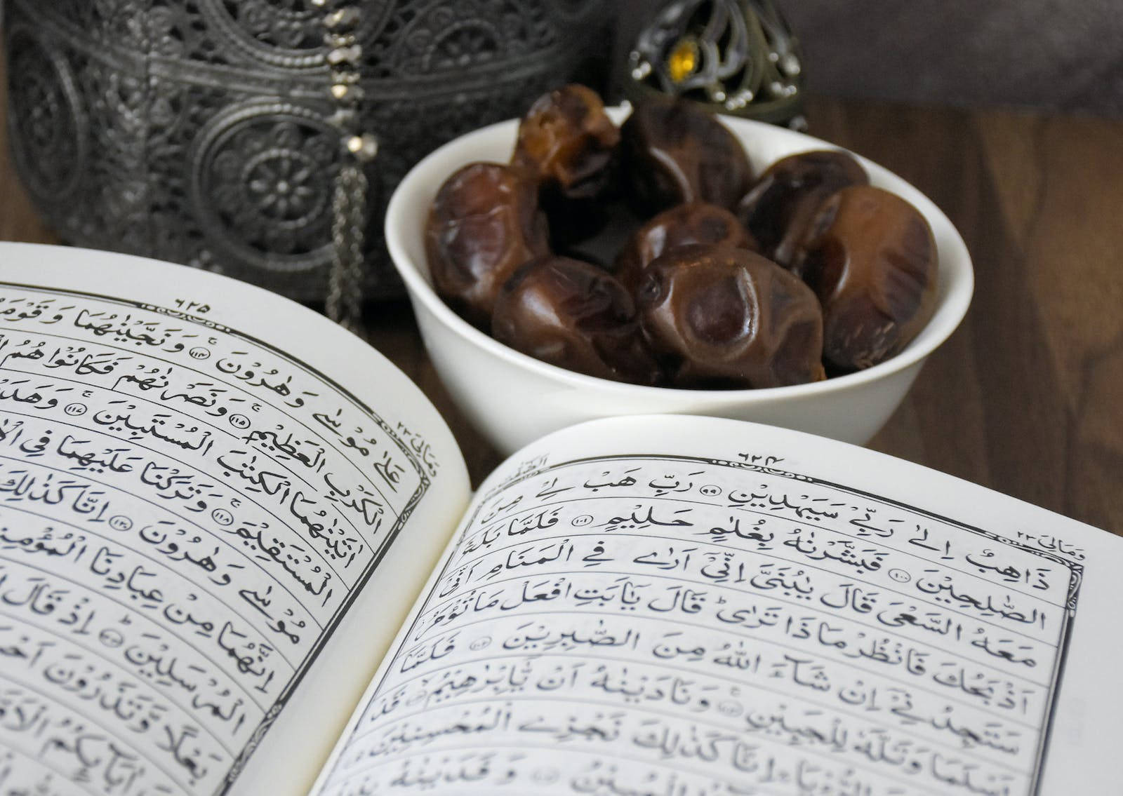 HD Islamic 桌布 壁紙 — 一本打開的古蘭經放在木質表面上，旁邊有一個白色碗裝著深色椰棗，背景有華麗的銀色物品。