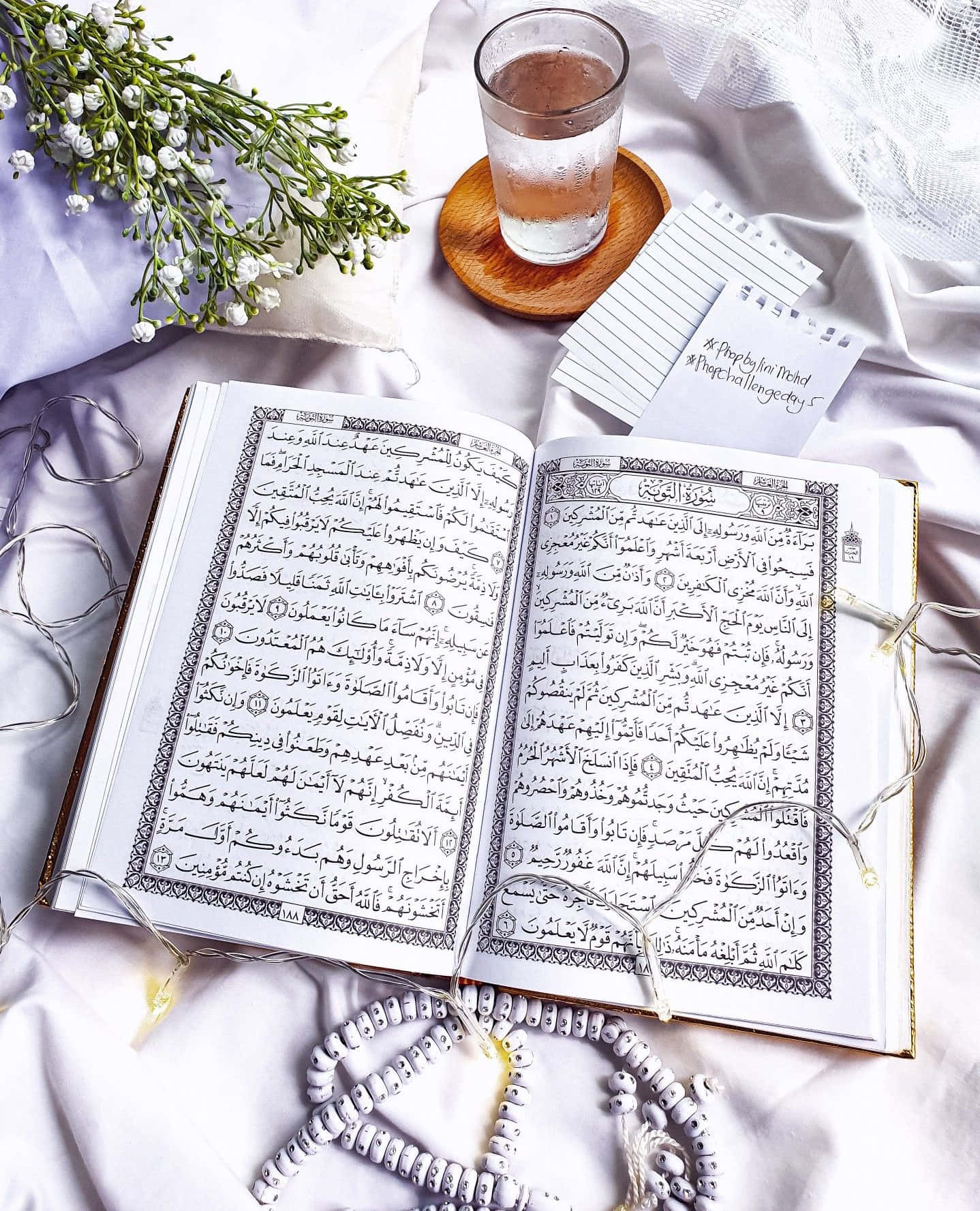 Quran 桌布 壁紙 — 一本打開的可蘭經放在柔軟的白色床罩上，旁邊有一杯水、祈禱珠和精緻的白色花朵。