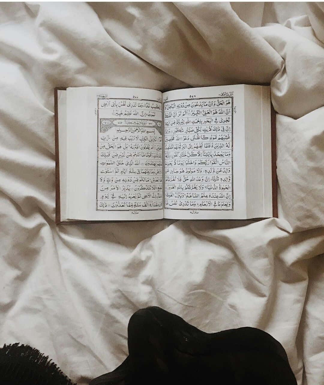 Quran 桌布 壁紙 — 一本打開的可蘭經放在皺巴巴的白色床單上，底部有一隻穿著黑色襪子的腳。