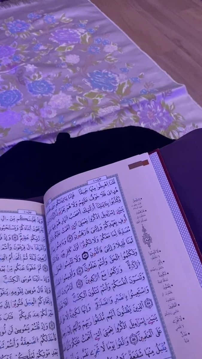 Quran 桌布 壁紙 — 一本打開的可蘭經放在花卉圖案的床罩上，柔和紫色燈光下。