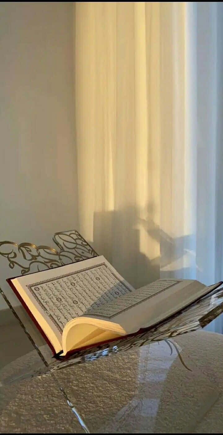 Quran 桌布 壁紙 — 一本打開的可蘭經放在華麗的支架上，沐浴在透過薄紗窗簾灑進的溫暖陽光中。