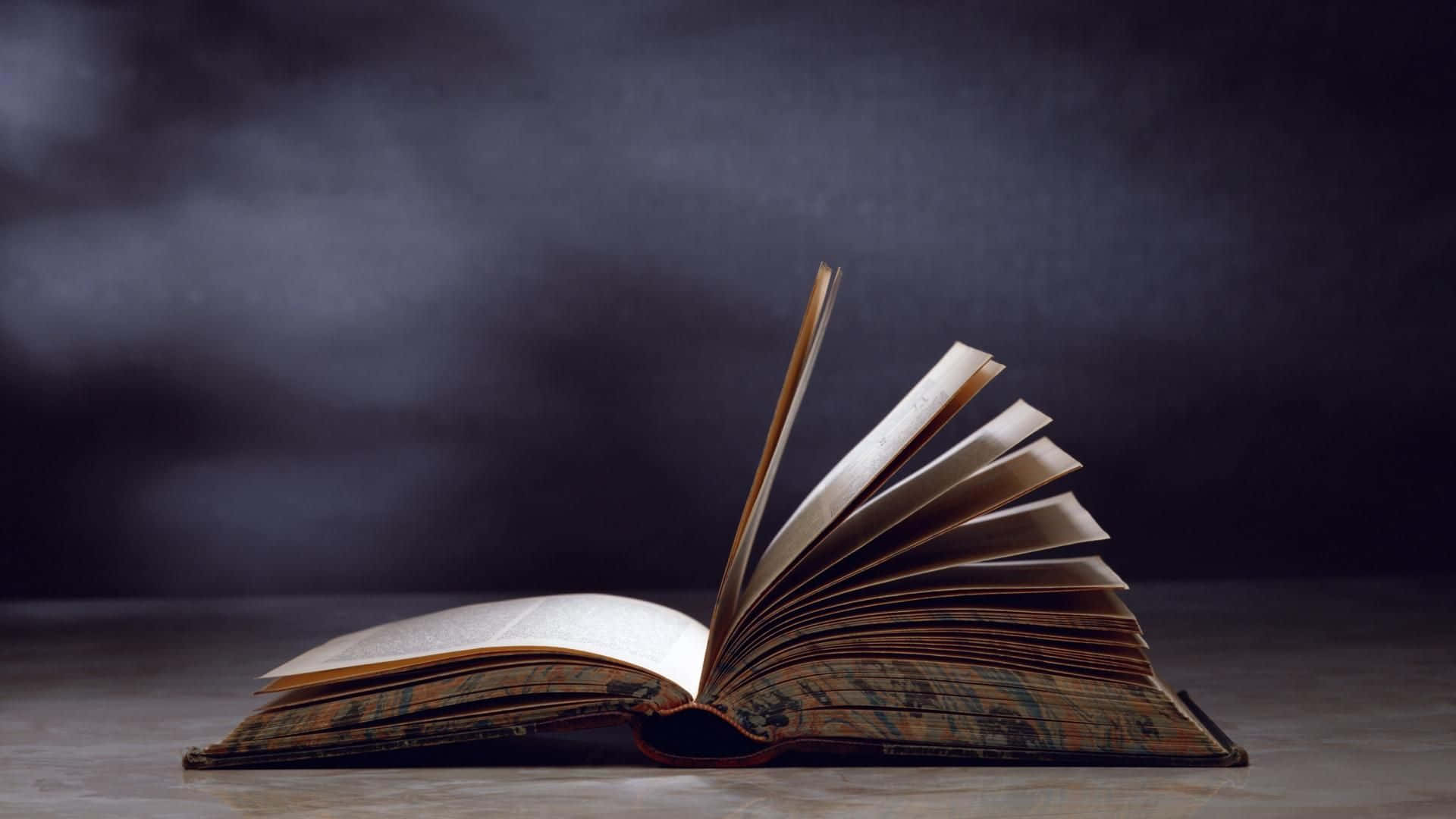 Educated 桌布 壁紙 — 一本打開的復古書，書頁如扇般展開，放在樸素的表面上，襯托著陰鬱的深色背景。